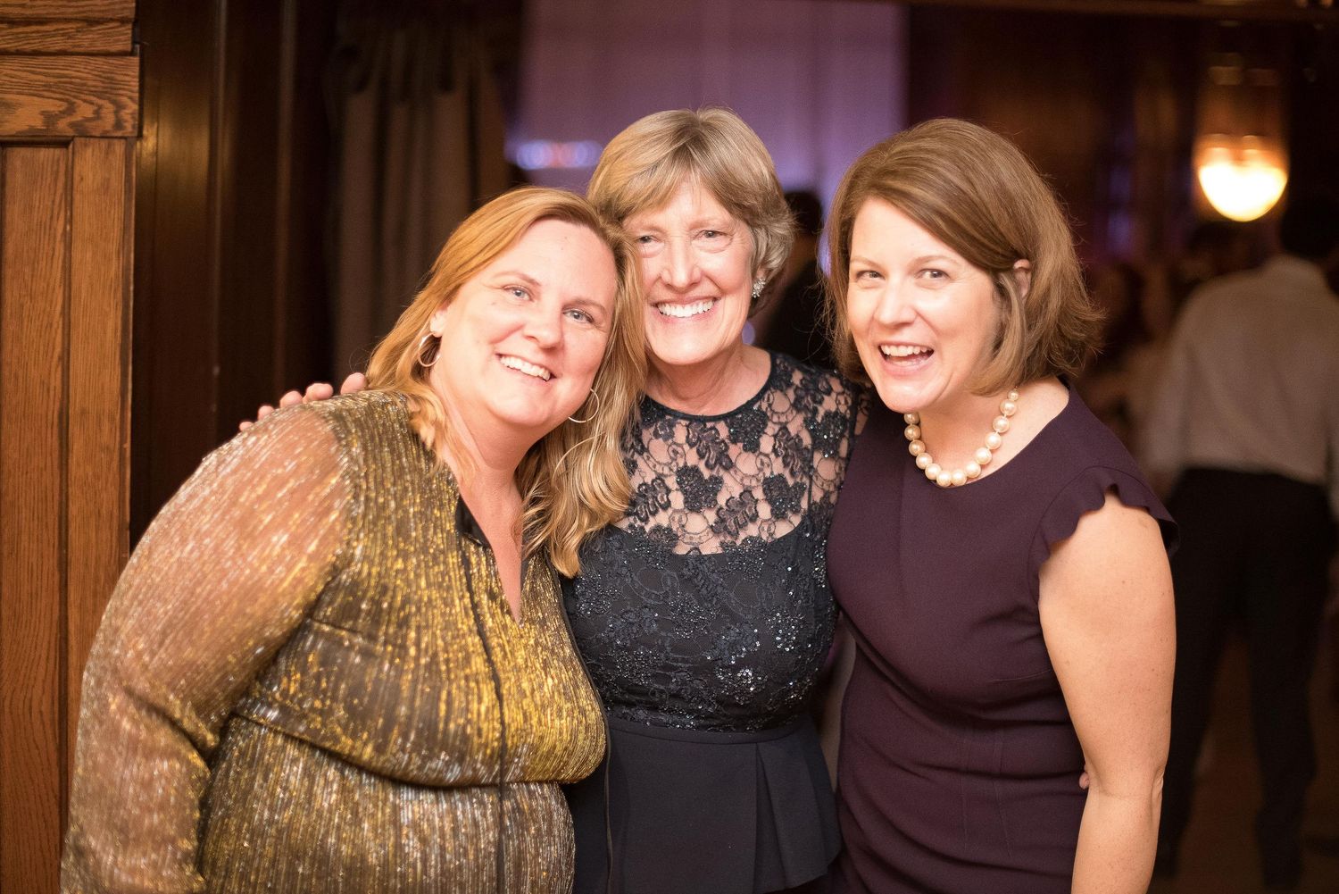 Three smiling women pose together at a formal event, wearing elegant attire in a warmly lit venue.