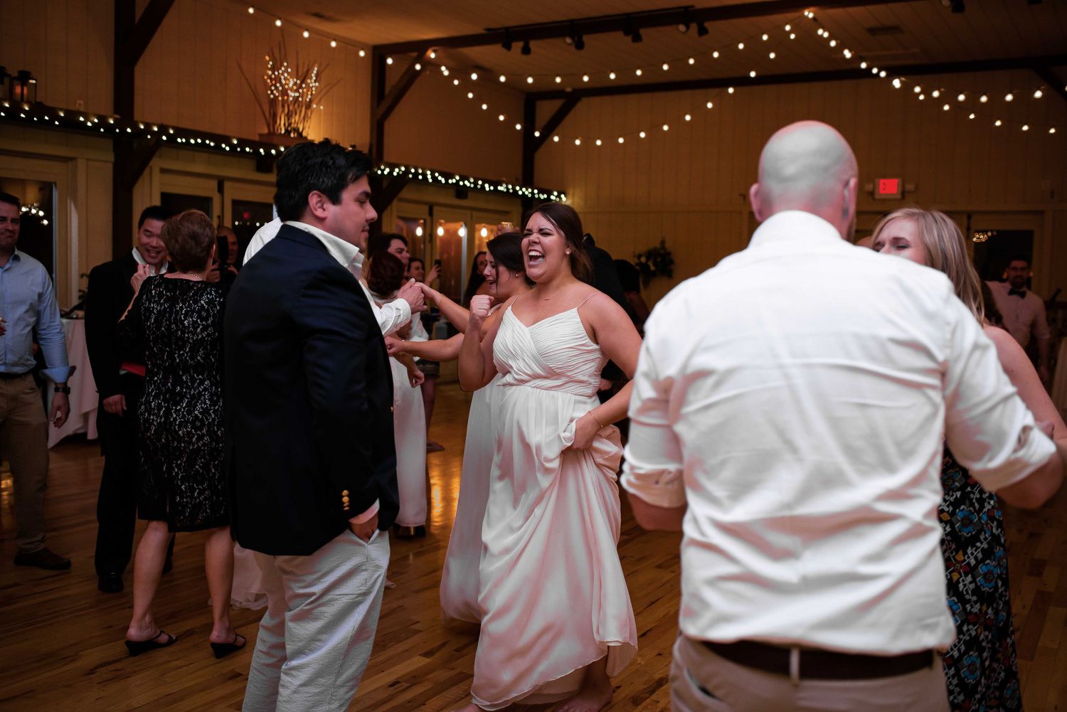 Guests laughing and dancing together at an elegant indoor wedding reception with string lights.