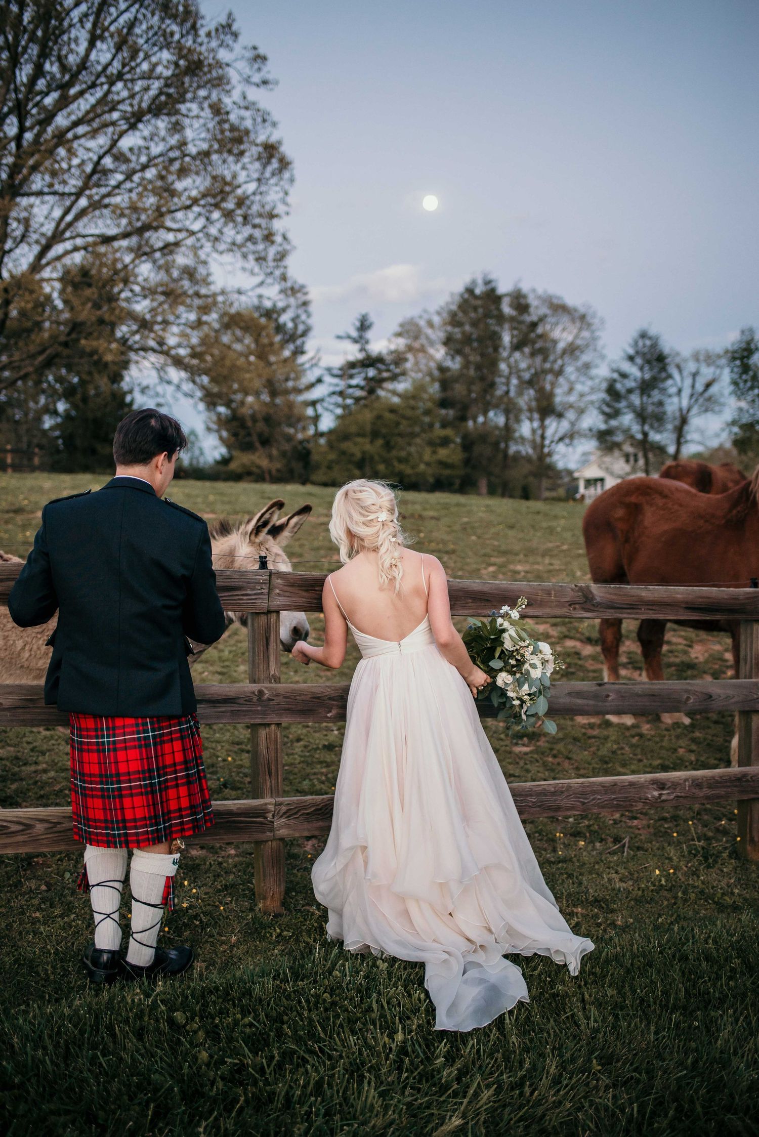 Bride in backless gown and groom in red tartan kilt stand at farm fence under full moon with horses.