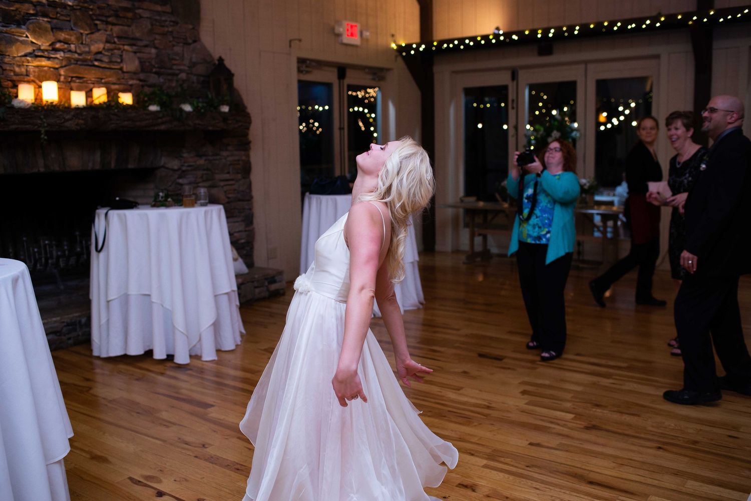 Bride in white gown dancing joyfully at indoor wedding reception with wood floors and stone fireplace.