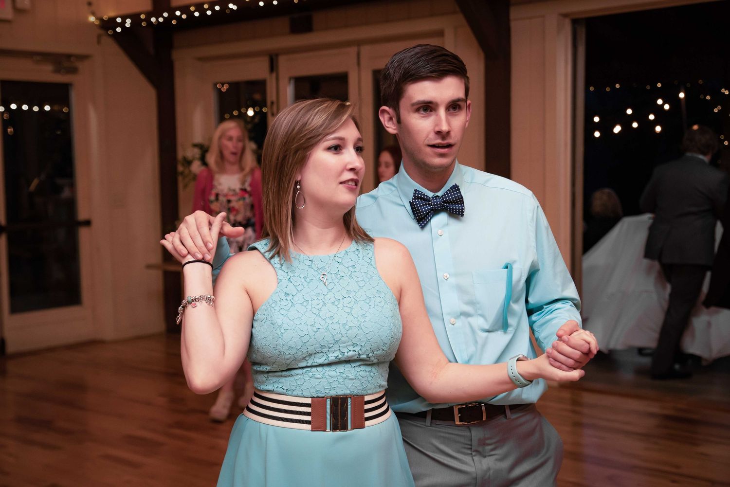 Couple dancing at wedding reception, woman in teal lace dress with striped belt, man in light blue shirt and bow tie.