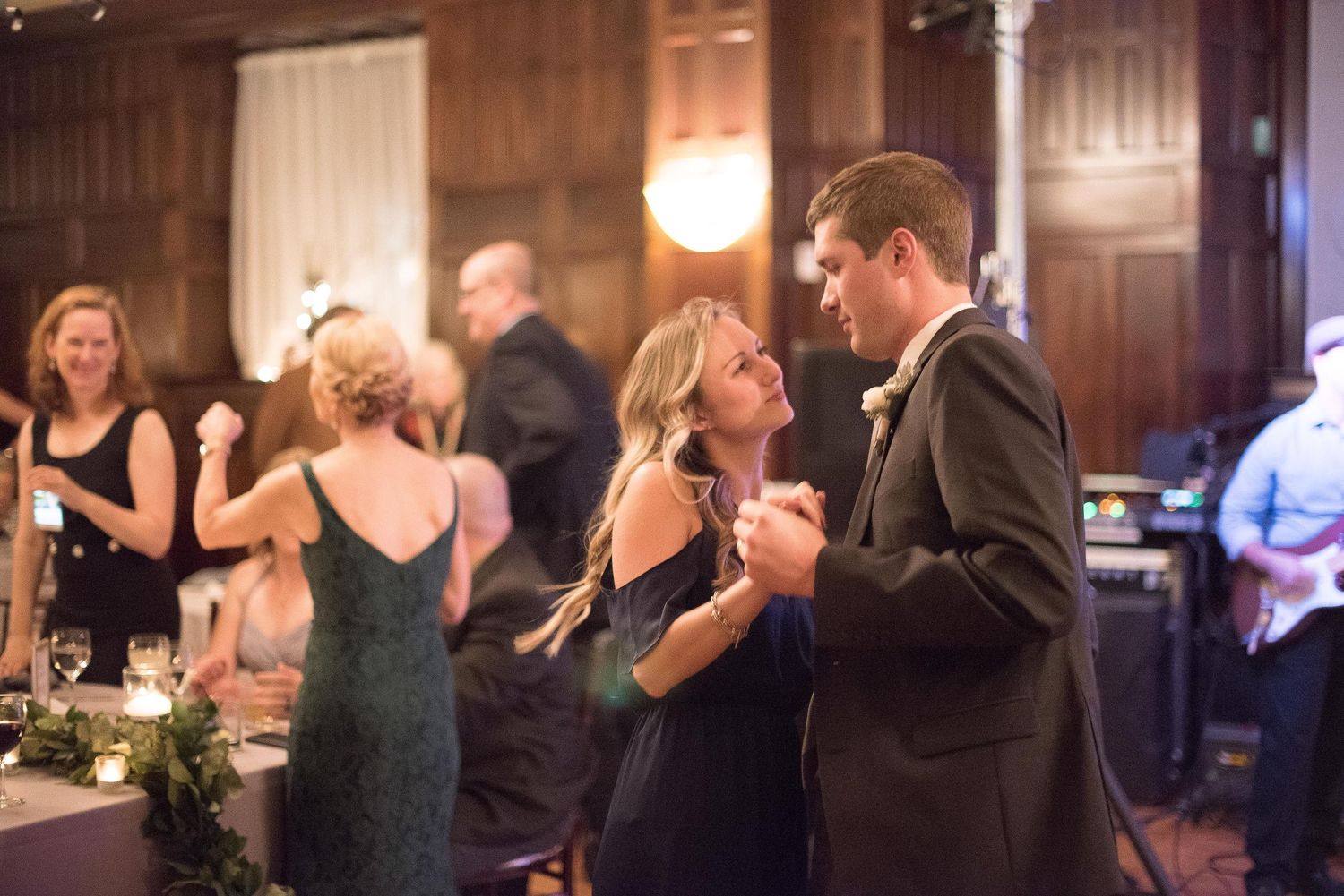 Couple sharing a romantic dance at an elegant wedding reception in a warmly lit venue with guests in background.