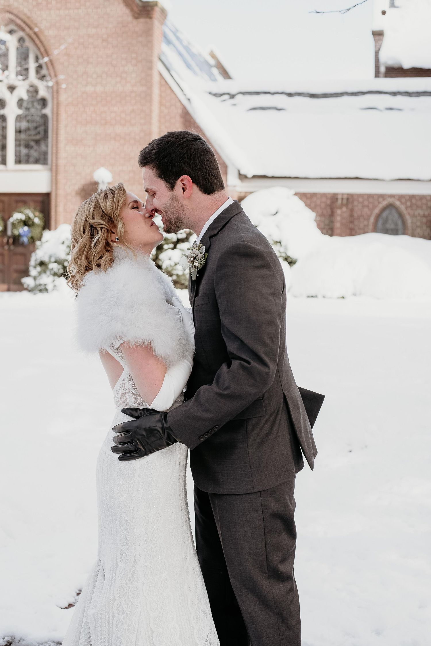 Bride and groom share a romantic moment in the snow outside a church, she wearing a white fur stole and lace gown.