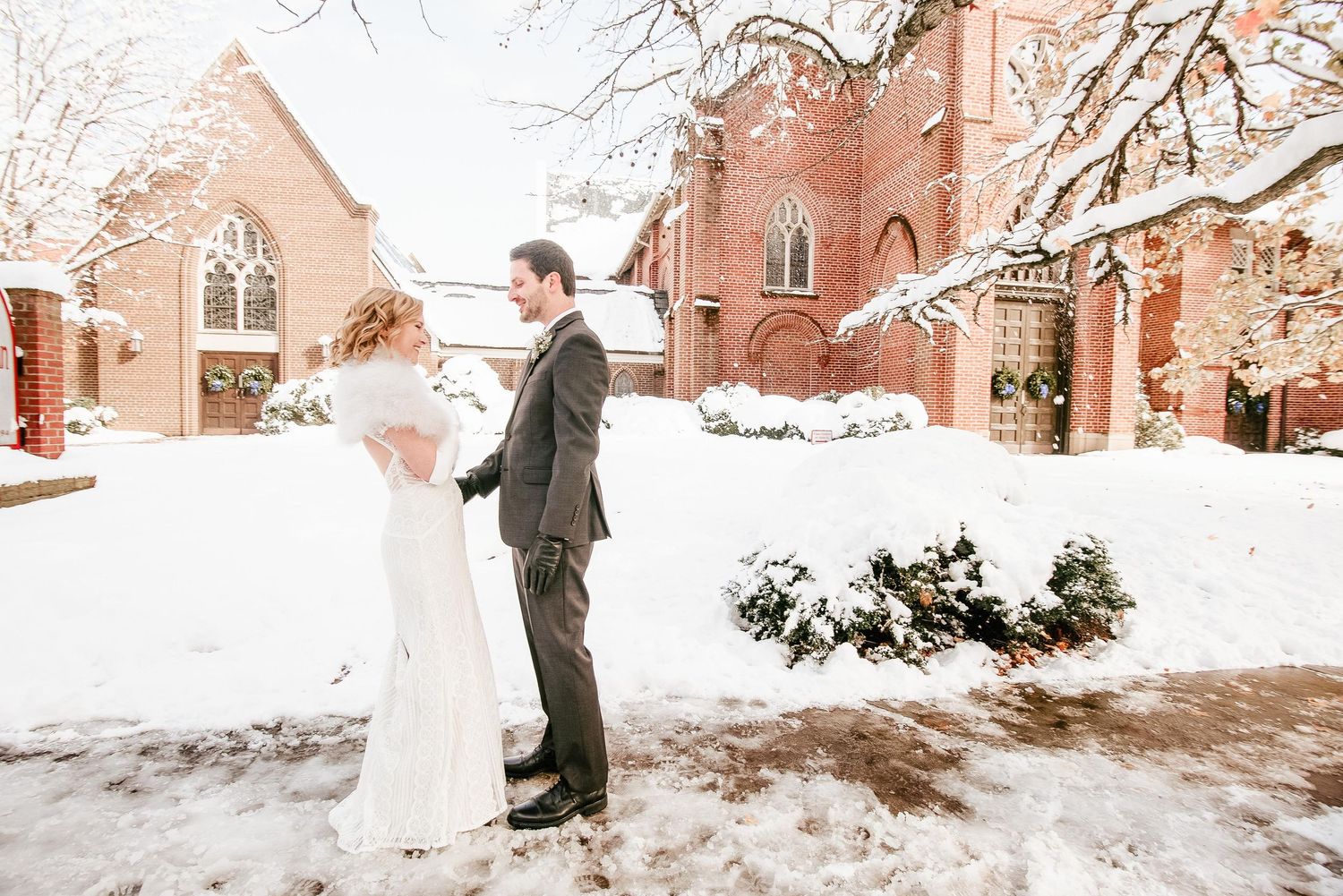 Bride and groom stand together in snow outside a red brick church on their winter wedding day.