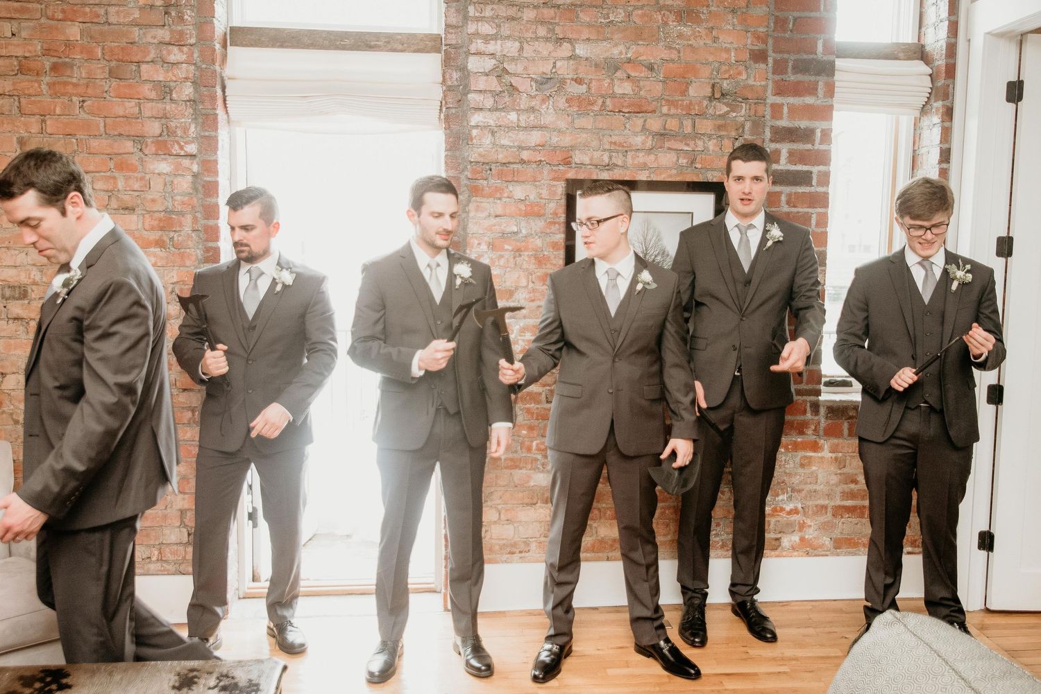 Groomsmen in gray suits adjusting ties and boutonnieres before a wedding ceremony in a brick-walled venue.