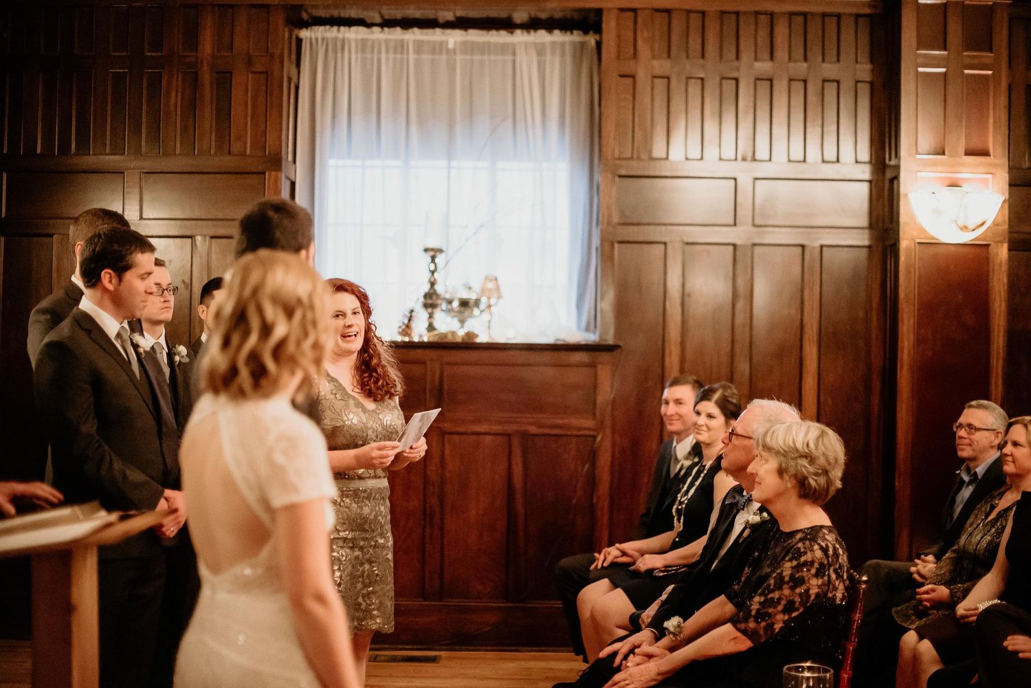 Wedding ceremony in wood-paneled room with bride, groom, and officiant reading vows before seated guests.