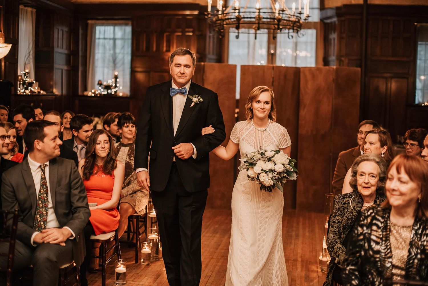 Bride walks down the aisle with escort in wood-paneled wedding venue filled with seated guests.