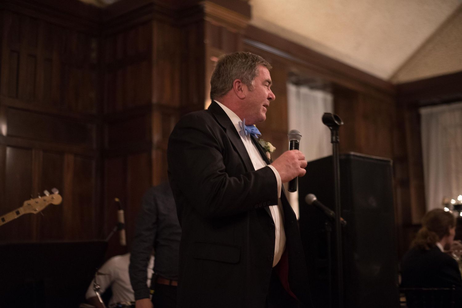 Man in tuxedo with bow tie speaking at microphone during formal wedding reception event.