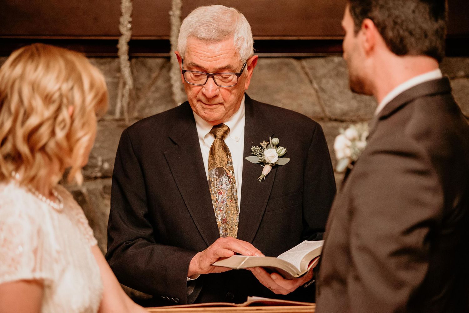 Elderly officiant reads from a book during an intimate wedding ceremony, wearing a boutonniere and gold tie.