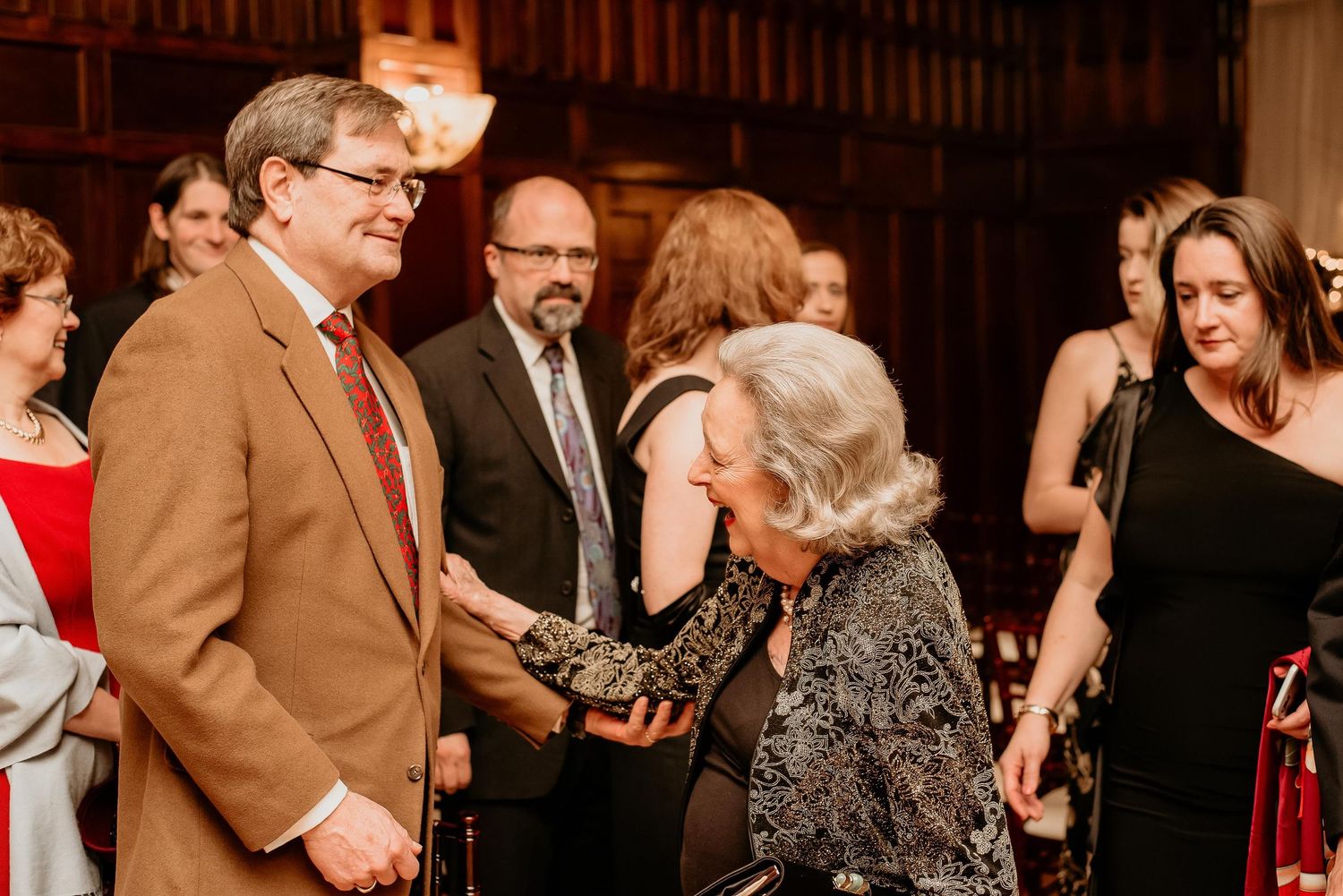 Guests mingling at an elegant formal event, with a man in a suit conversing with an elderly woman in a decorated venue.