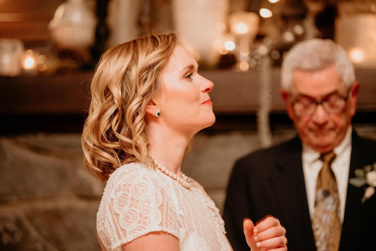 Bride in white lace dress and pearl necklace smiling at wedding reception with warm candlelit background.