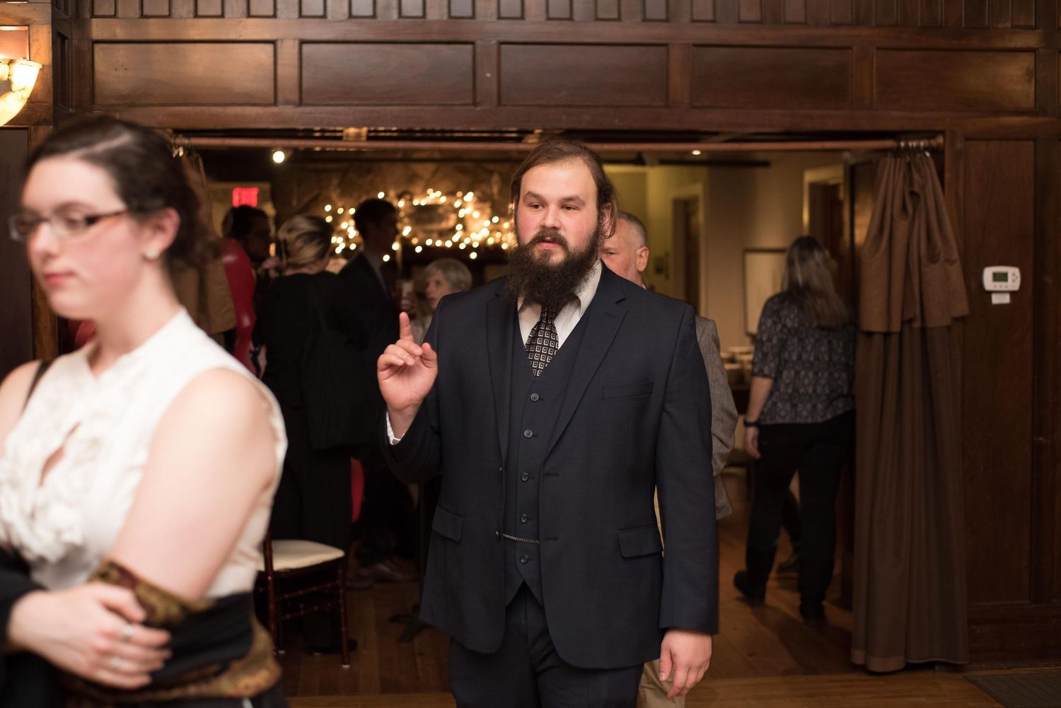 Bearded man in dark suit gesturing with finger at elegant indoor event with warm string lights in background.