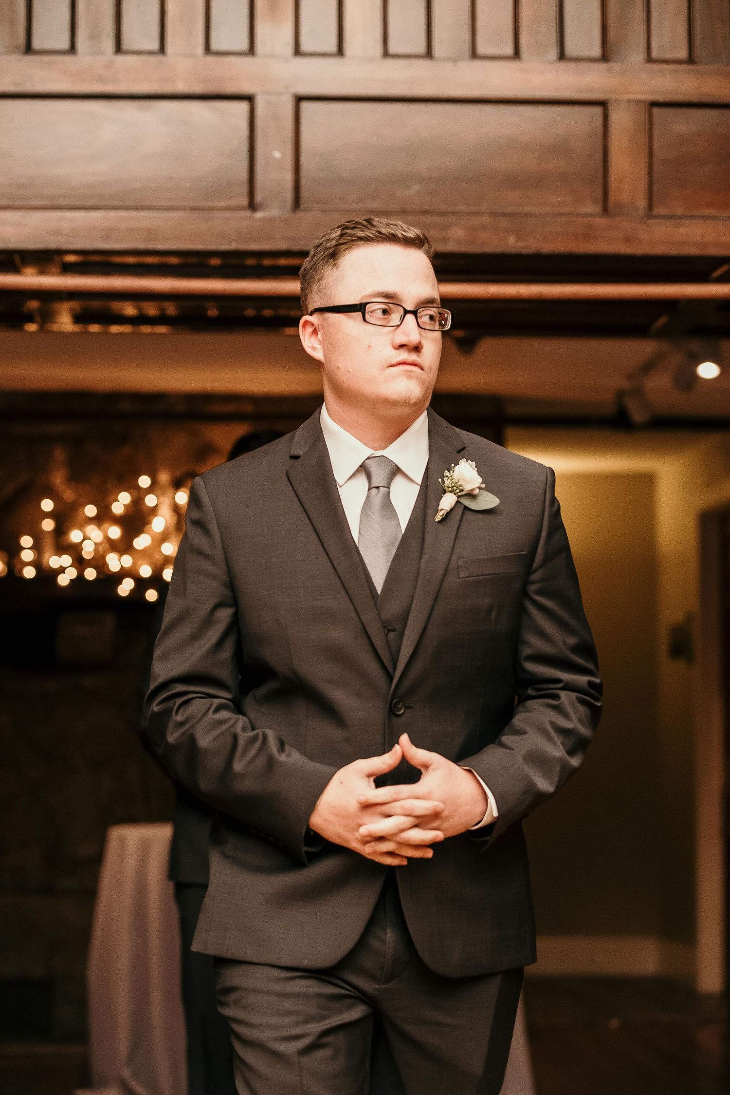 Groom in dark suit with boutonniere and gray tie stands elegantly at wedding reception venue with warm lights.