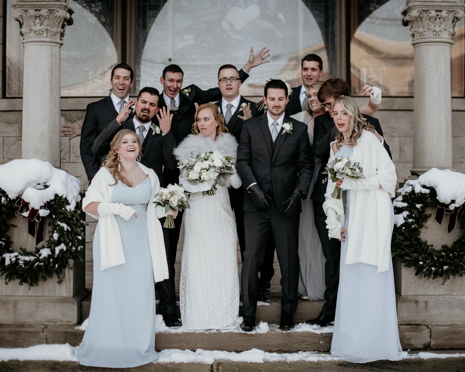 Winter wedding party poses on snowy steps, bride and groom centered with bridesmaids in white and groomsmen in dark suits.