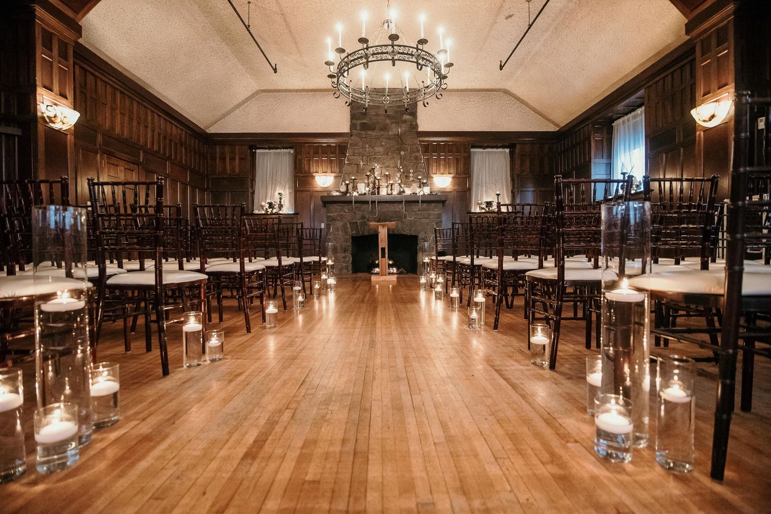 Elegant wedding ceremony setup with dark wood chairs, candlelit aisle, hardwood floors, and grand chandeliers in a classic ballroom.