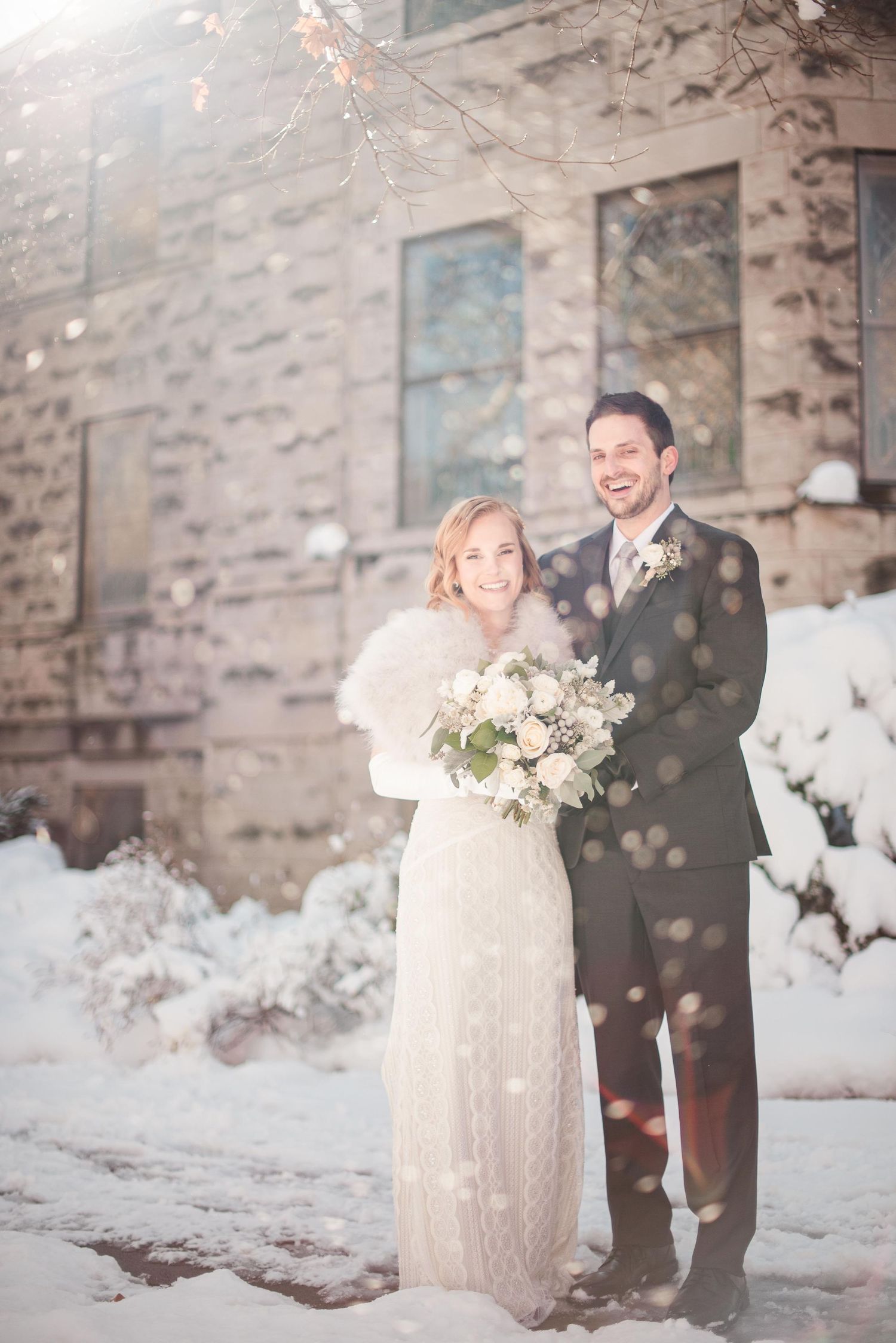 Bride and groom smiling together in snow outside stone building, bride holding white bouquet wearing fur wrap over wedding dress.