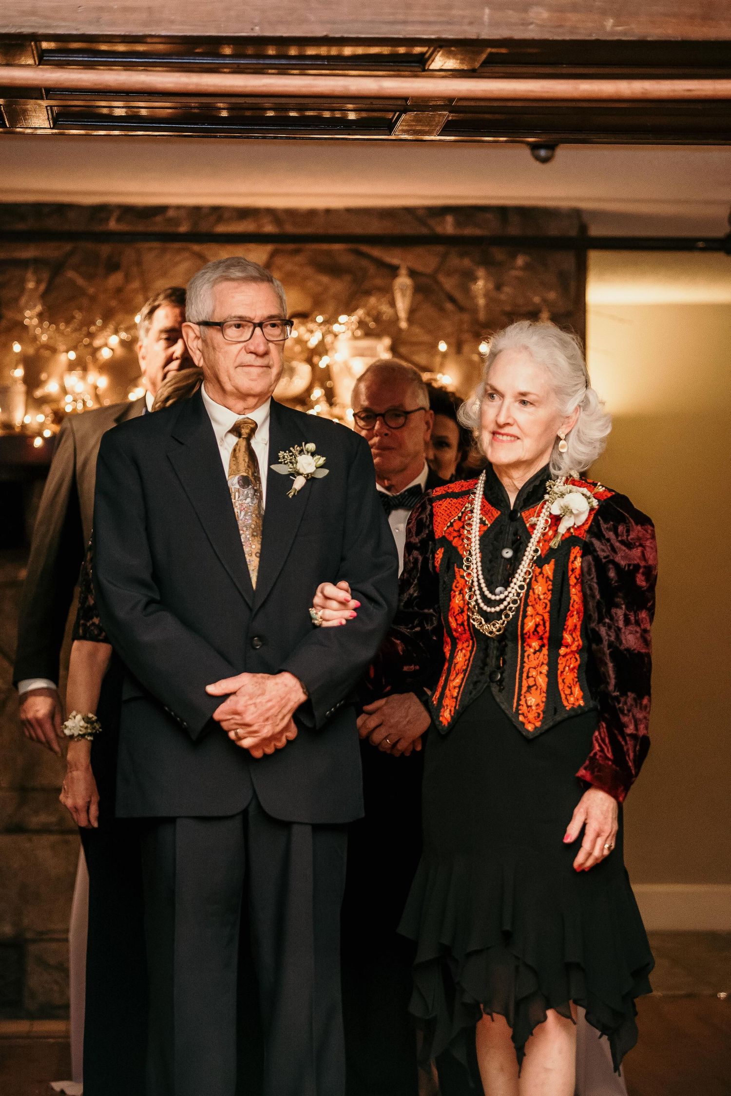 Elegant elderly couple in formal attire walking together at a wedding reception with warm fairy lights in background.