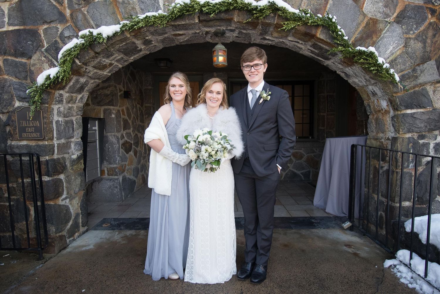 Bride in white gown with bouquet poses with bridesmaid and groom under stone arch in winter wedding.