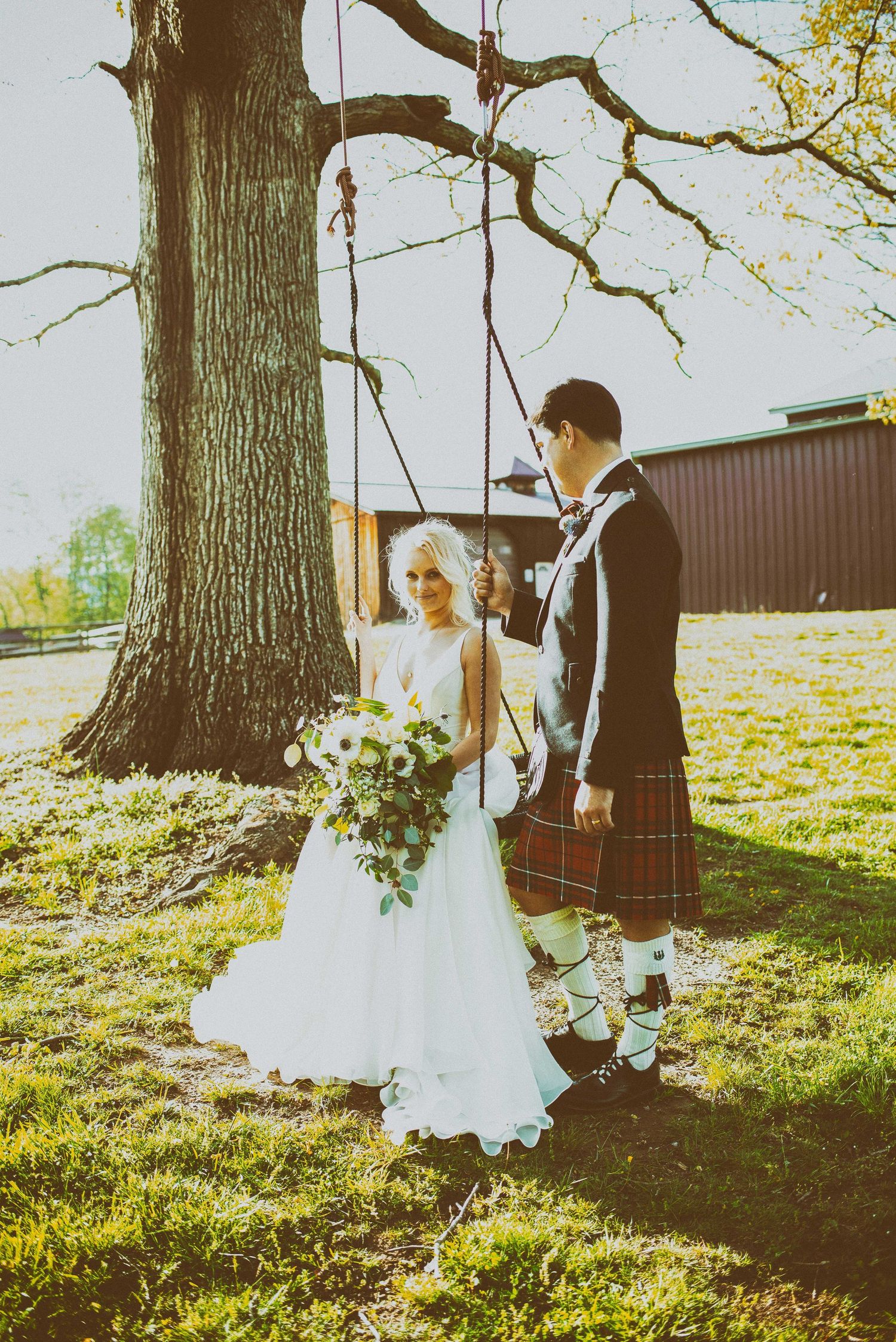 Bride in white gown holds bouquet beside groom in Scottish kilt near a tree swing at outdoor wedding.