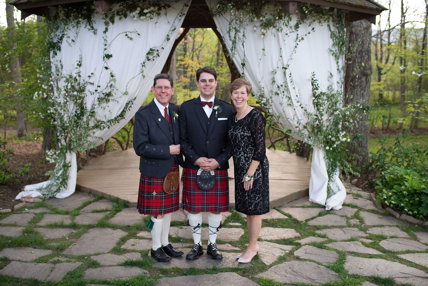 Three people pose in front of a floral gazebo, two men in kilts and a woman in a black dress at an outdoor wedding.