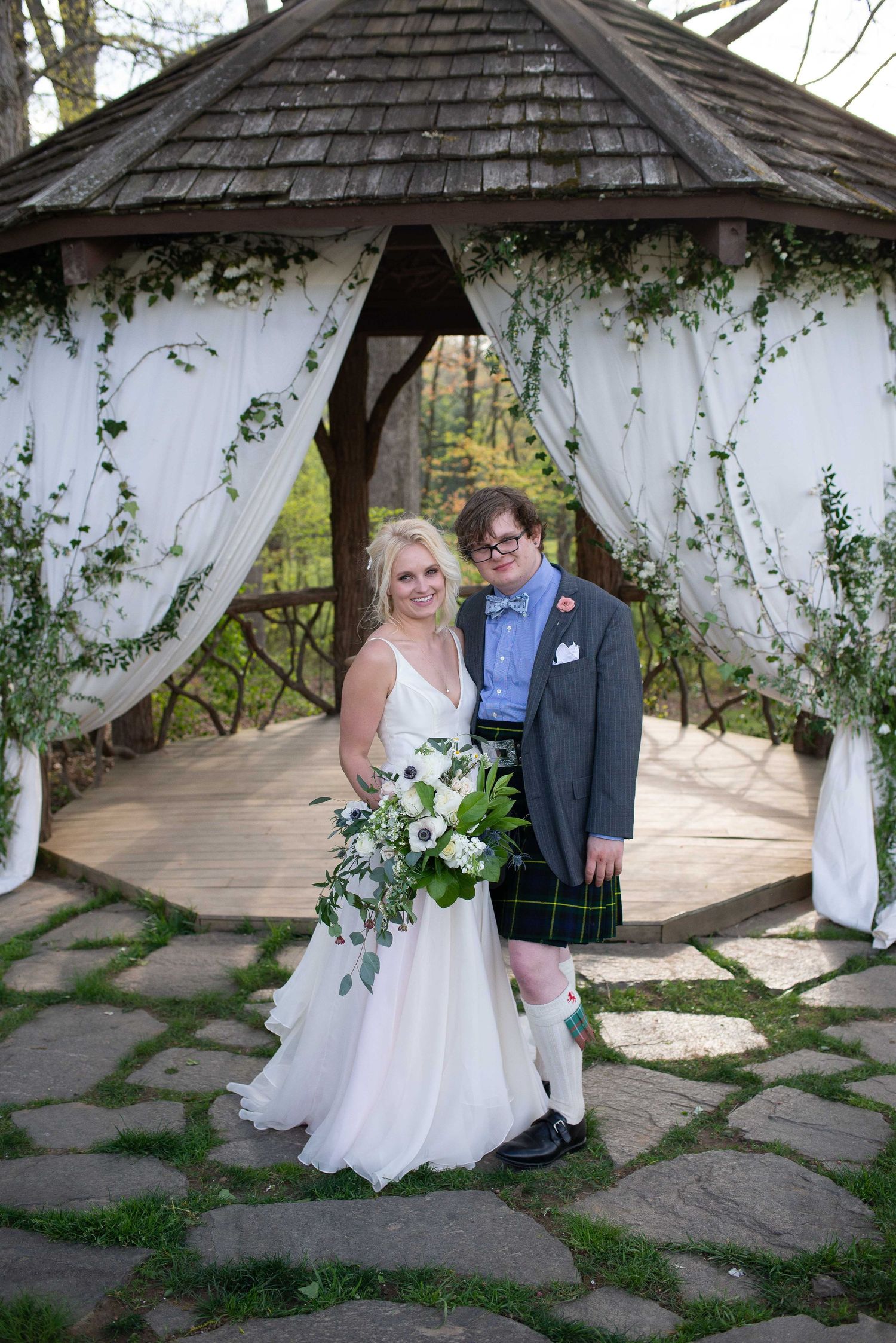 Bride and groom pose under decorated gazebo with white drapes and greenery, groom wearing tartan kilt at outdoor wedding.