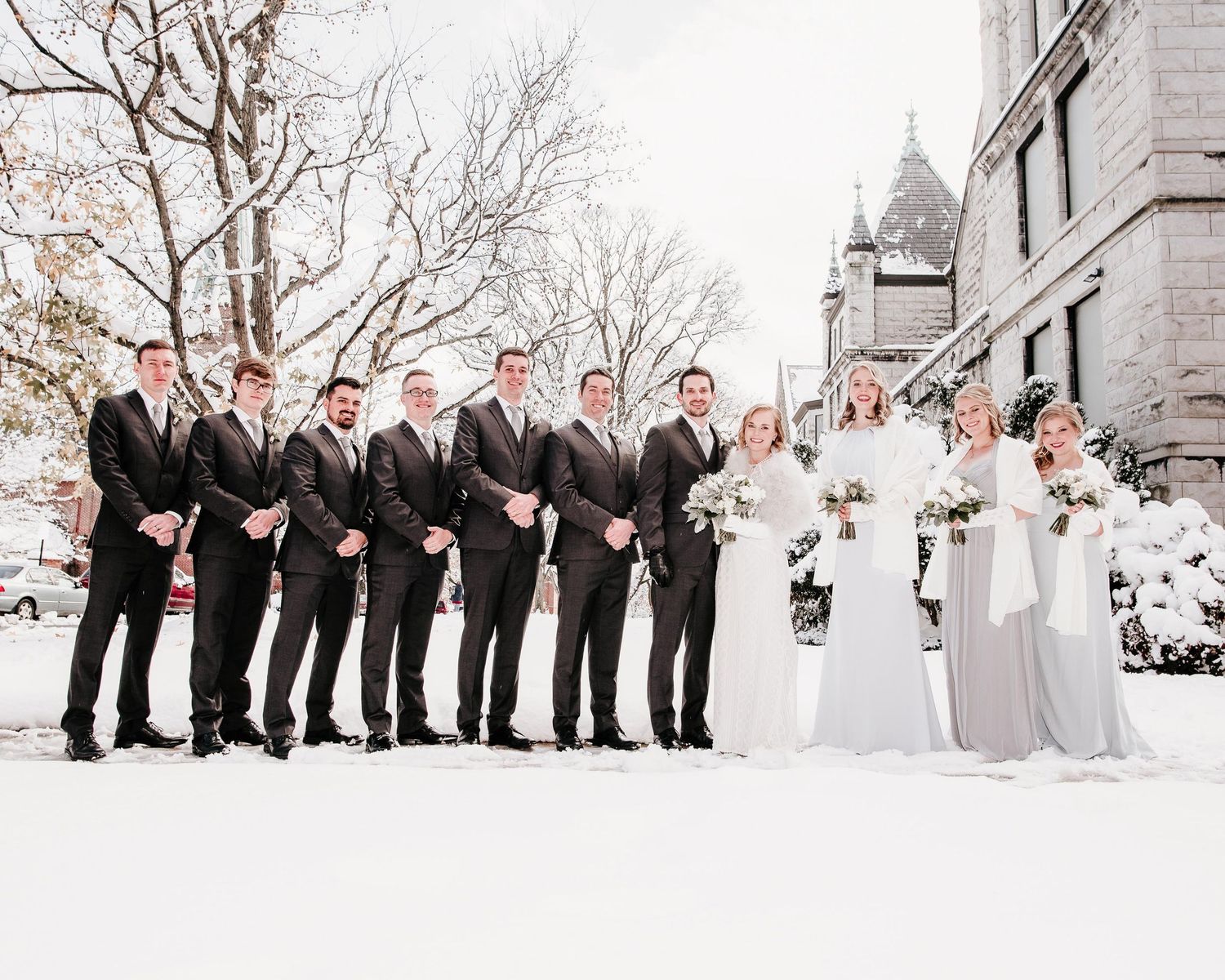 Wedding party posing in snow, groomsmen in black suits and bridesmaids in gray gowns holding white bouquets.