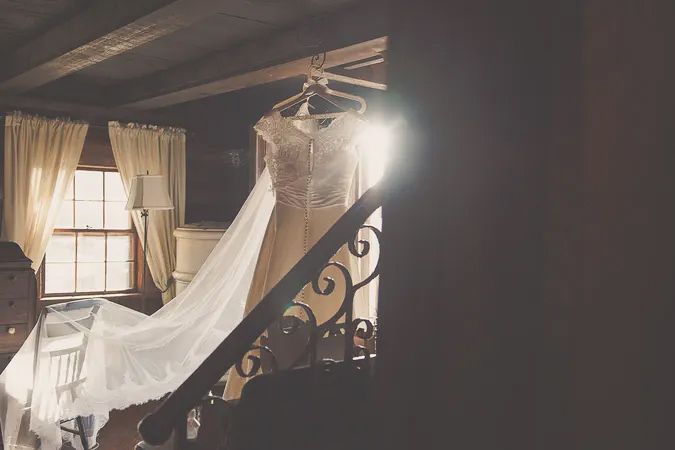 Elegant wedding dress hanging on ornate staircase railing in dimly lit rustic venue with curtained windows.