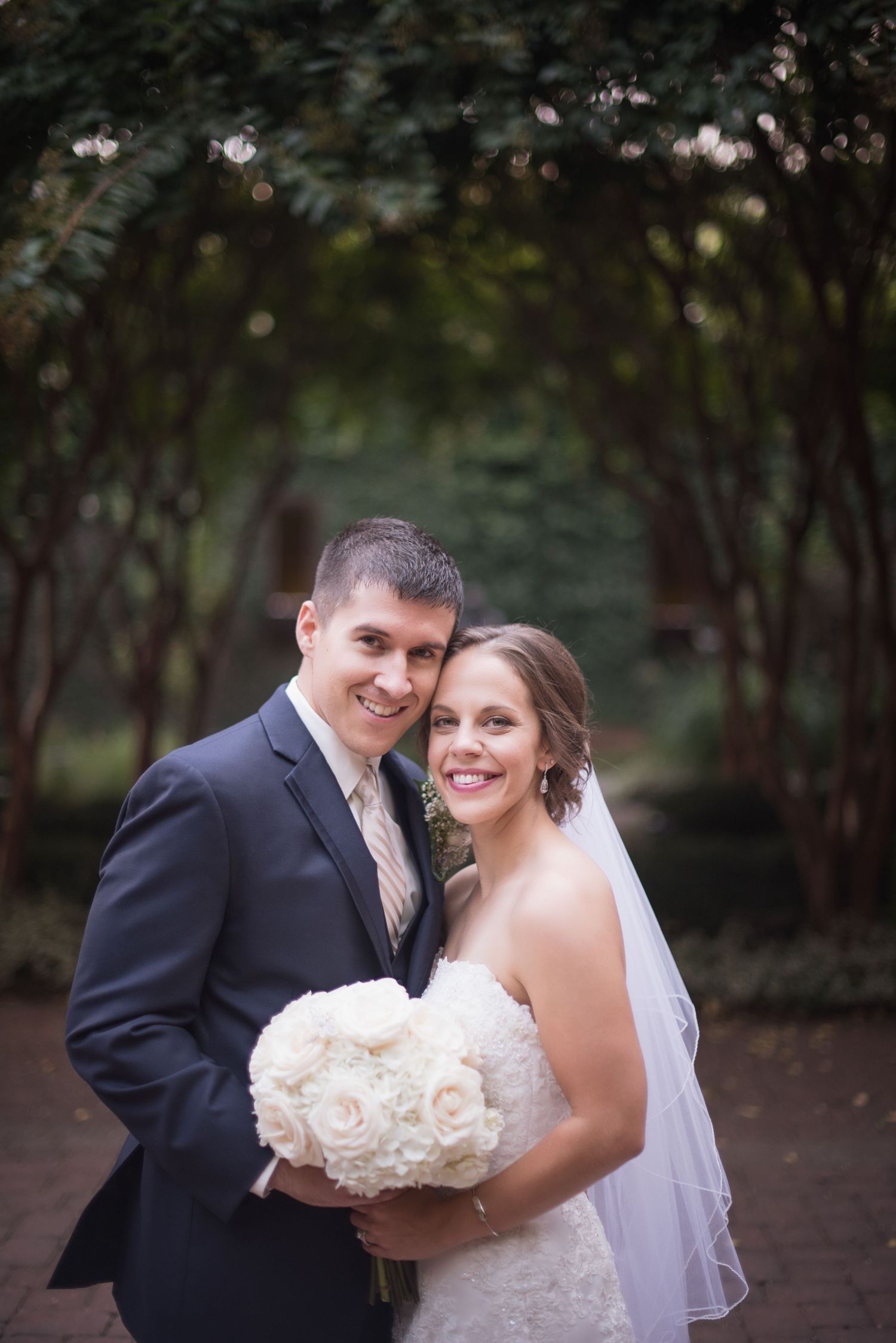 Happy bride and groom pose together outdoors under green tree archway, bride holding white rose bouquet.