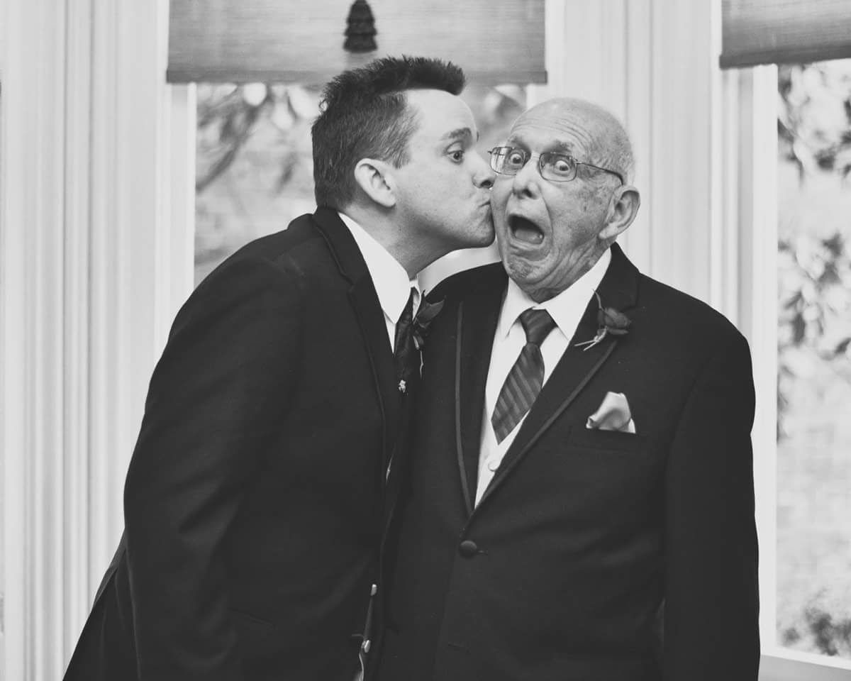 Groom kissing elderly man on cheek with surprised expression in black and white photo.