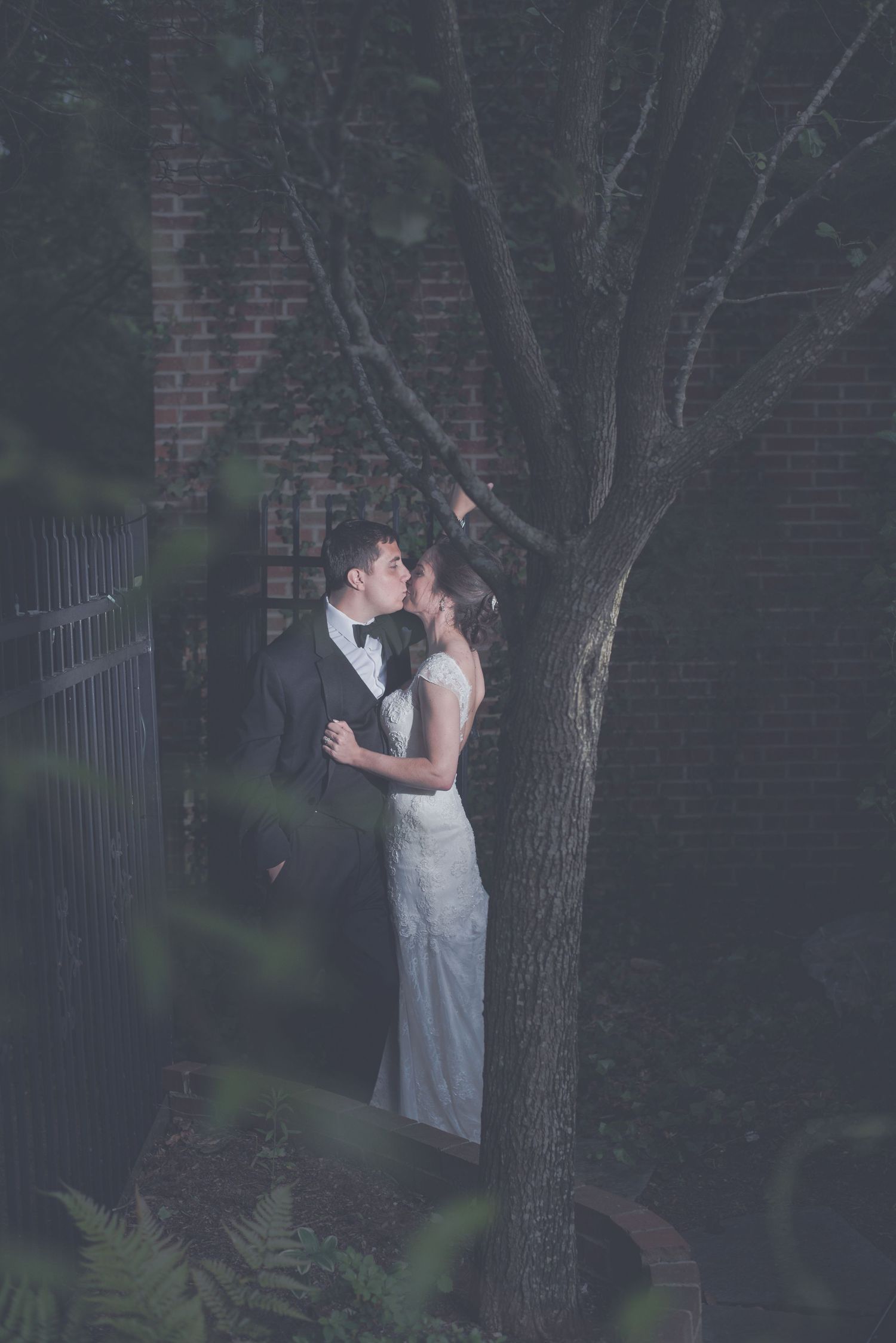 Bride and groom share a romantic kiss beneath a bare tree against a brick wall in a moody, dimly lit outdoor setting.