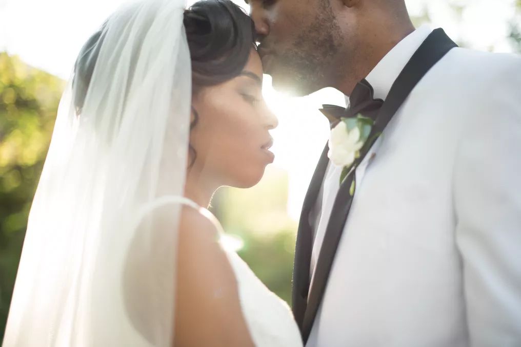 A bride and groom share a romantic kiss outdoors, with the bride wearing a white veil on their wedding day.