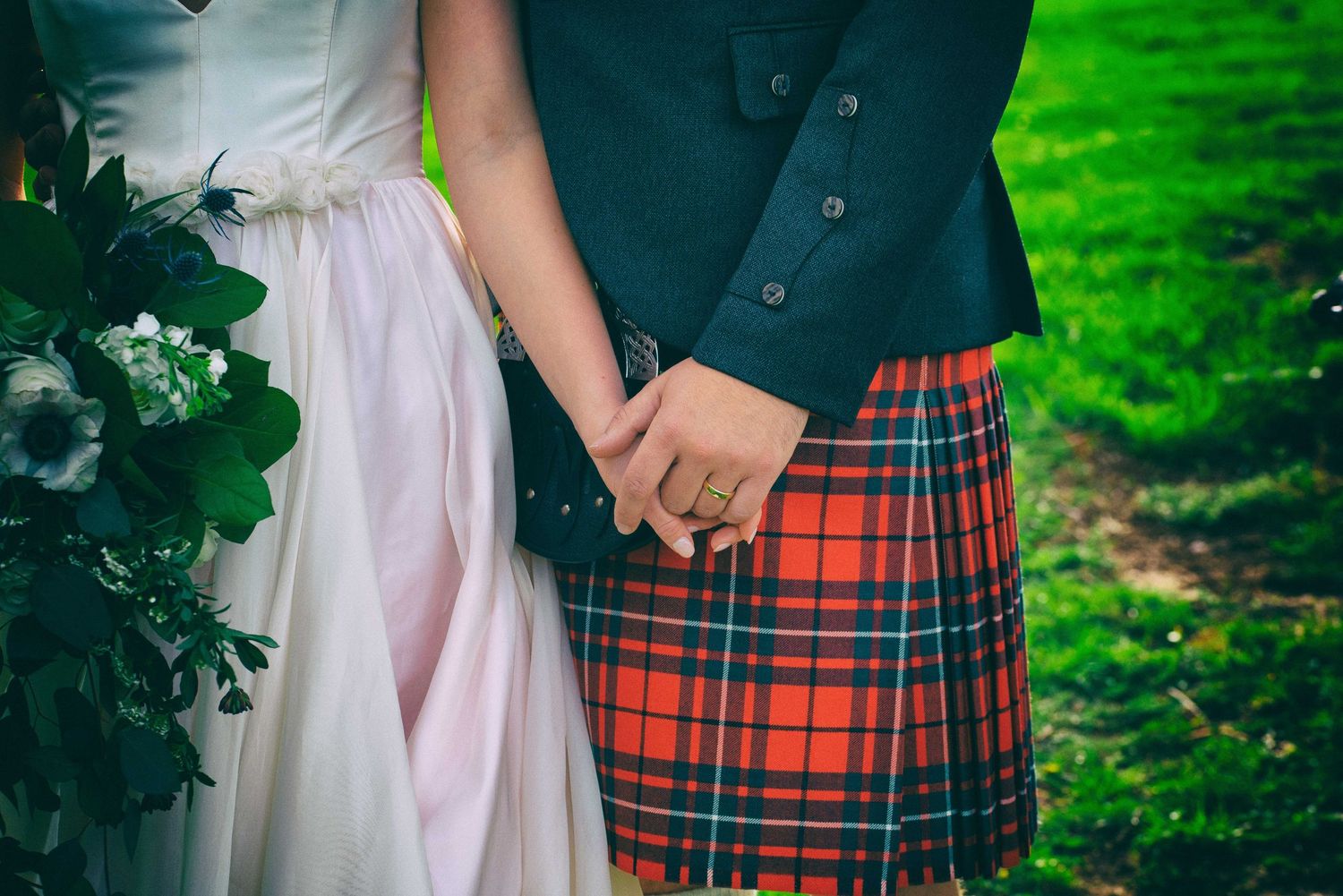 Bride and groom in red tartan kilt holding hands outdoors, bride holding green bouquet in white wedding dress.