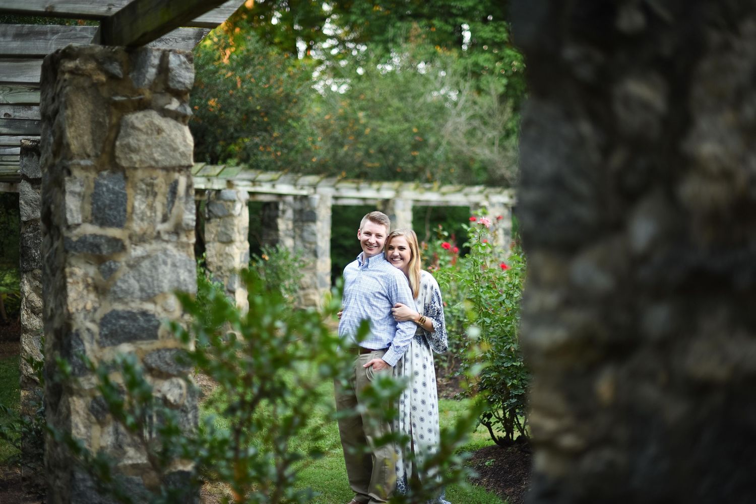 Couple embracing in a lush garden framed by rustic stone pillars and a wooden pergola with blooming flowers.
