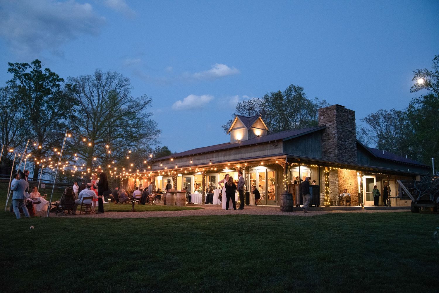 Rustic wedding venue with string lights and guests gathered outside at dusk.