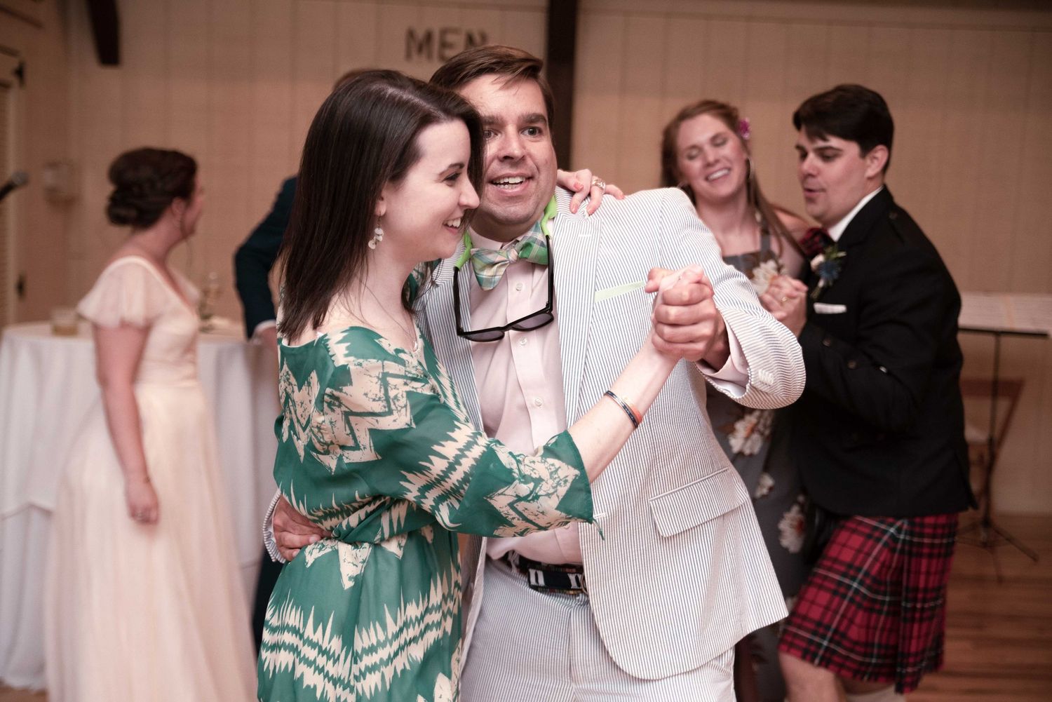 Couple dancing at wedding reception, man in white suit with bow tie, woman in green patterned dress, guests dancing behind them.