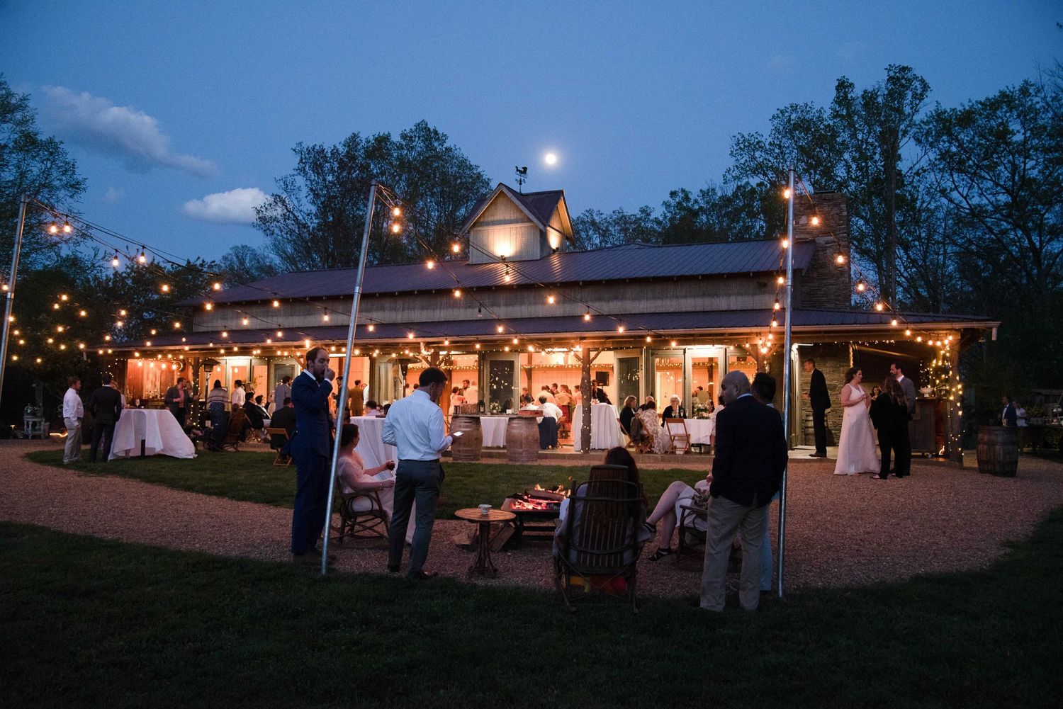Outdoor evening wedding reception with string lights adorning a white barn venue, guests mingling on the lawn at dusk.