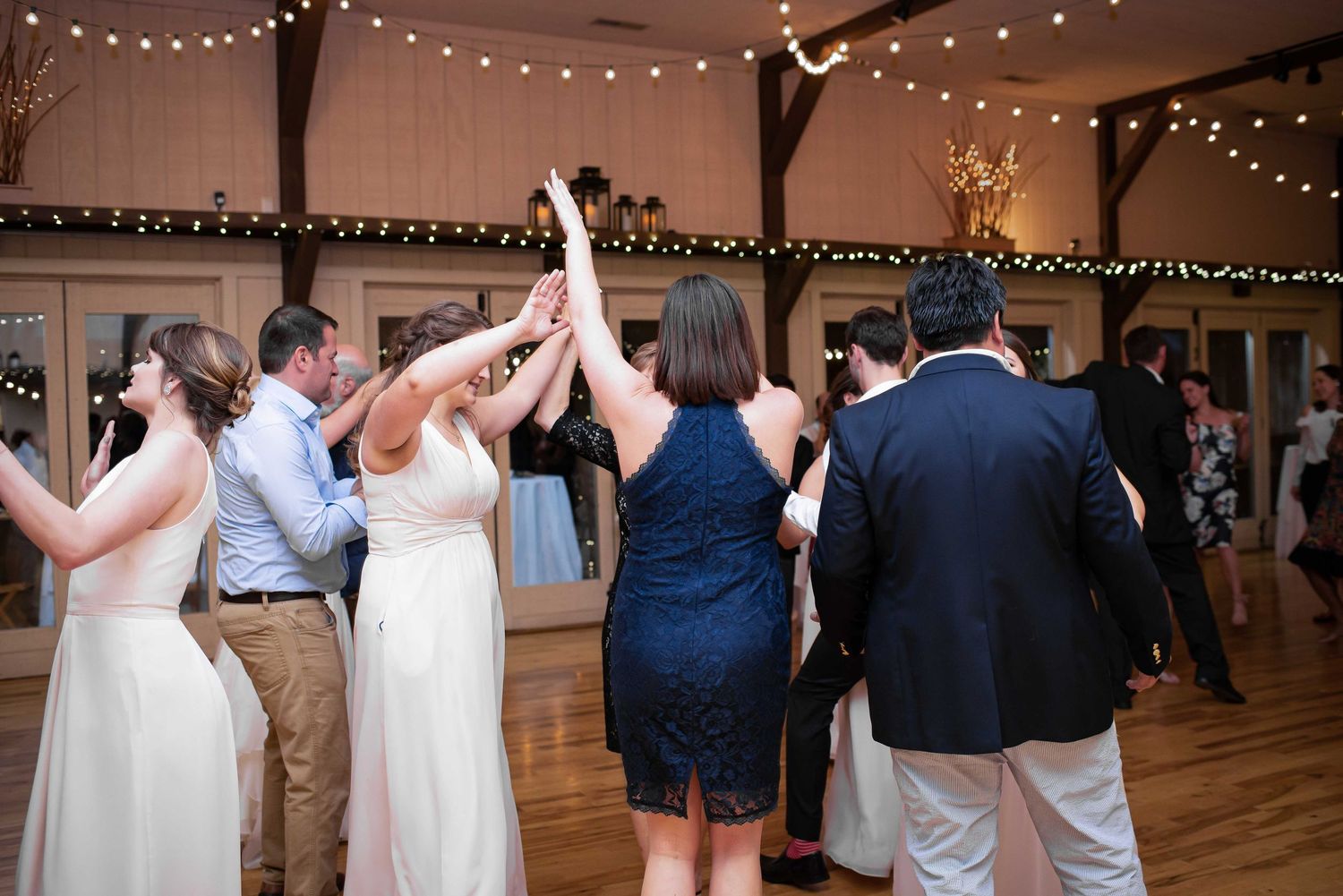 Wedding guests dancing together in a warmly lit reception hall decorated with string lights and rustic floral arrangements.