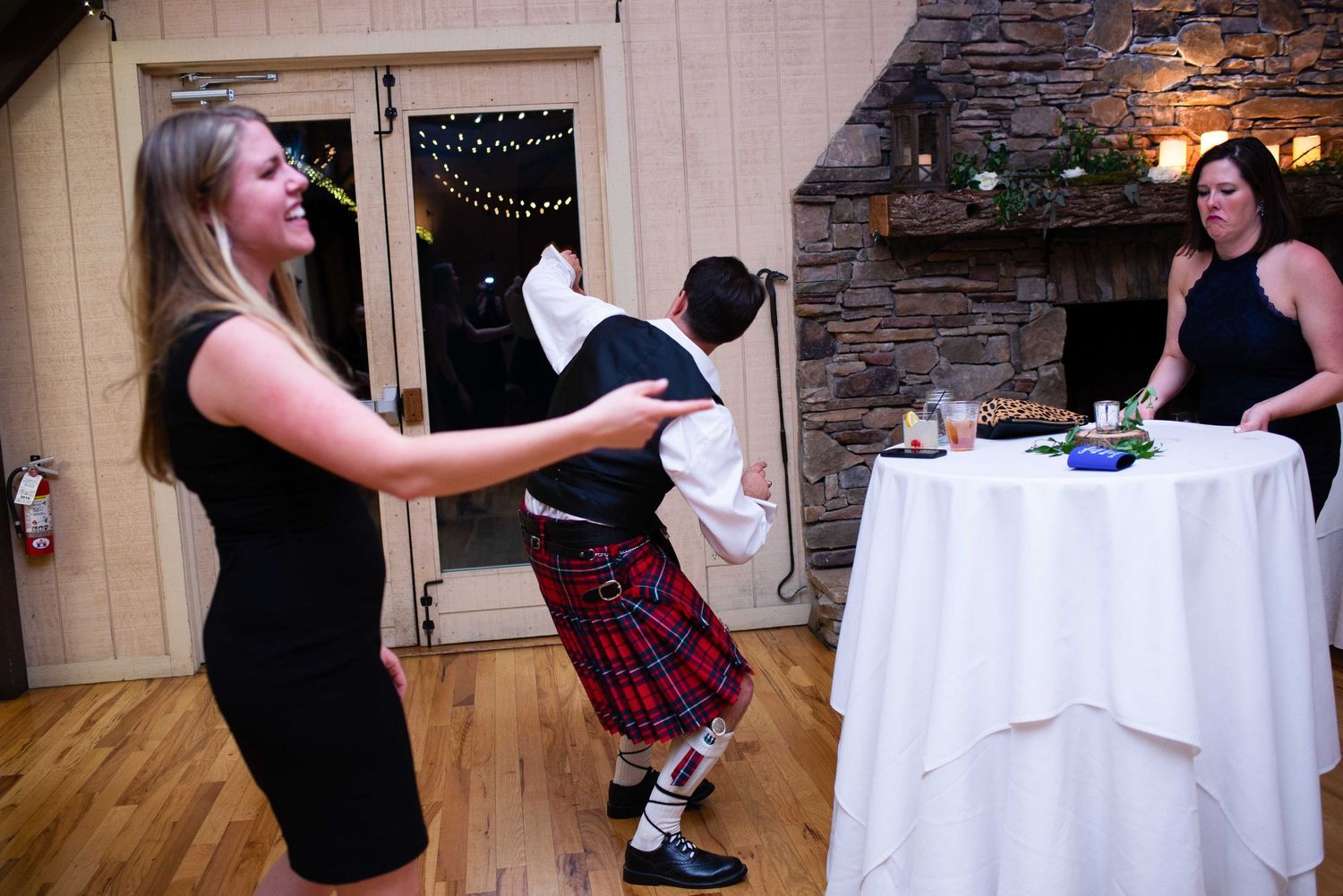 Wedding reception scene with woman in black dress dancing with man in traditional Scottish kilt near stone fireplace.