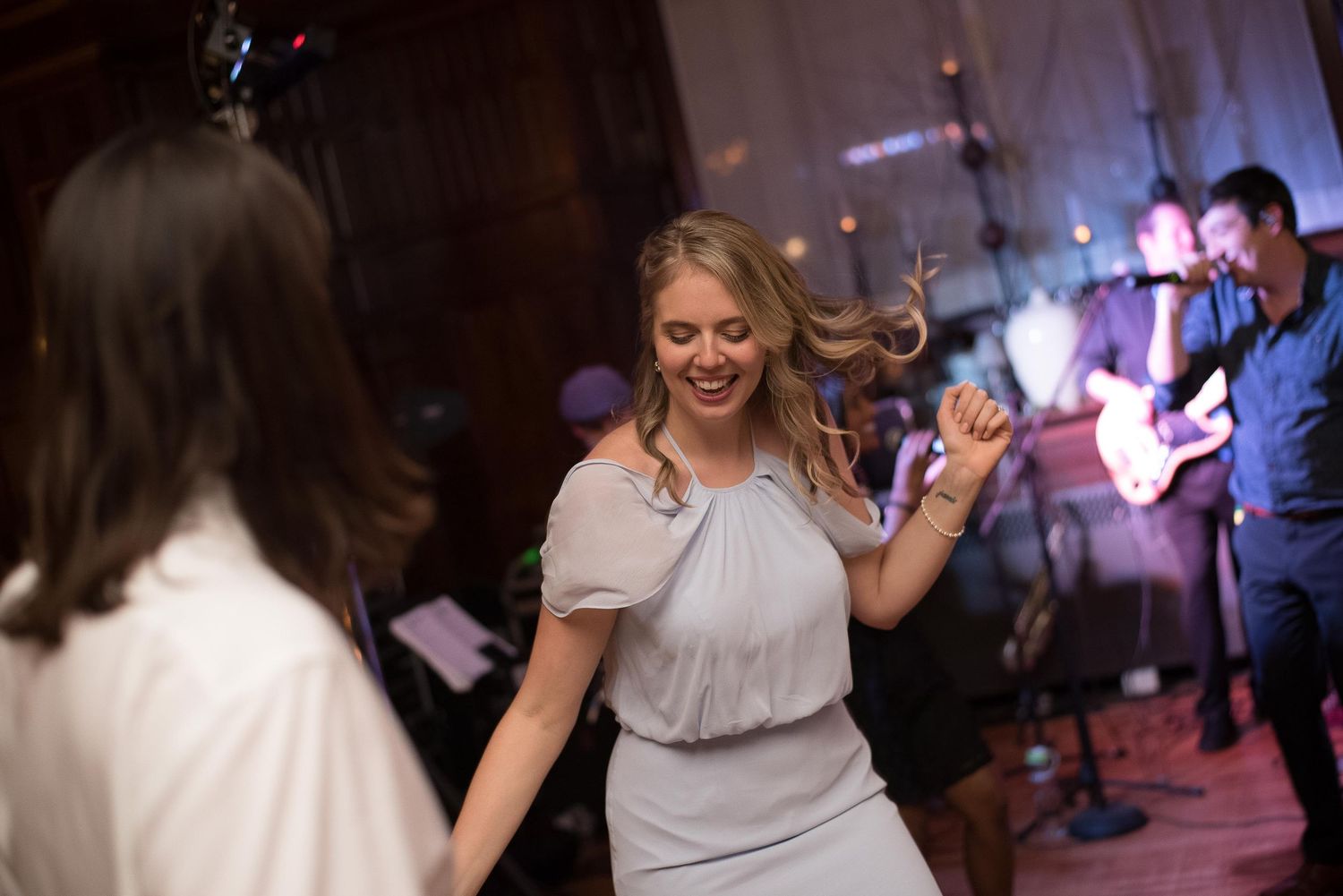 Woman in gray dress dancing joyfully at a wedding reception with a live band performing in the background.