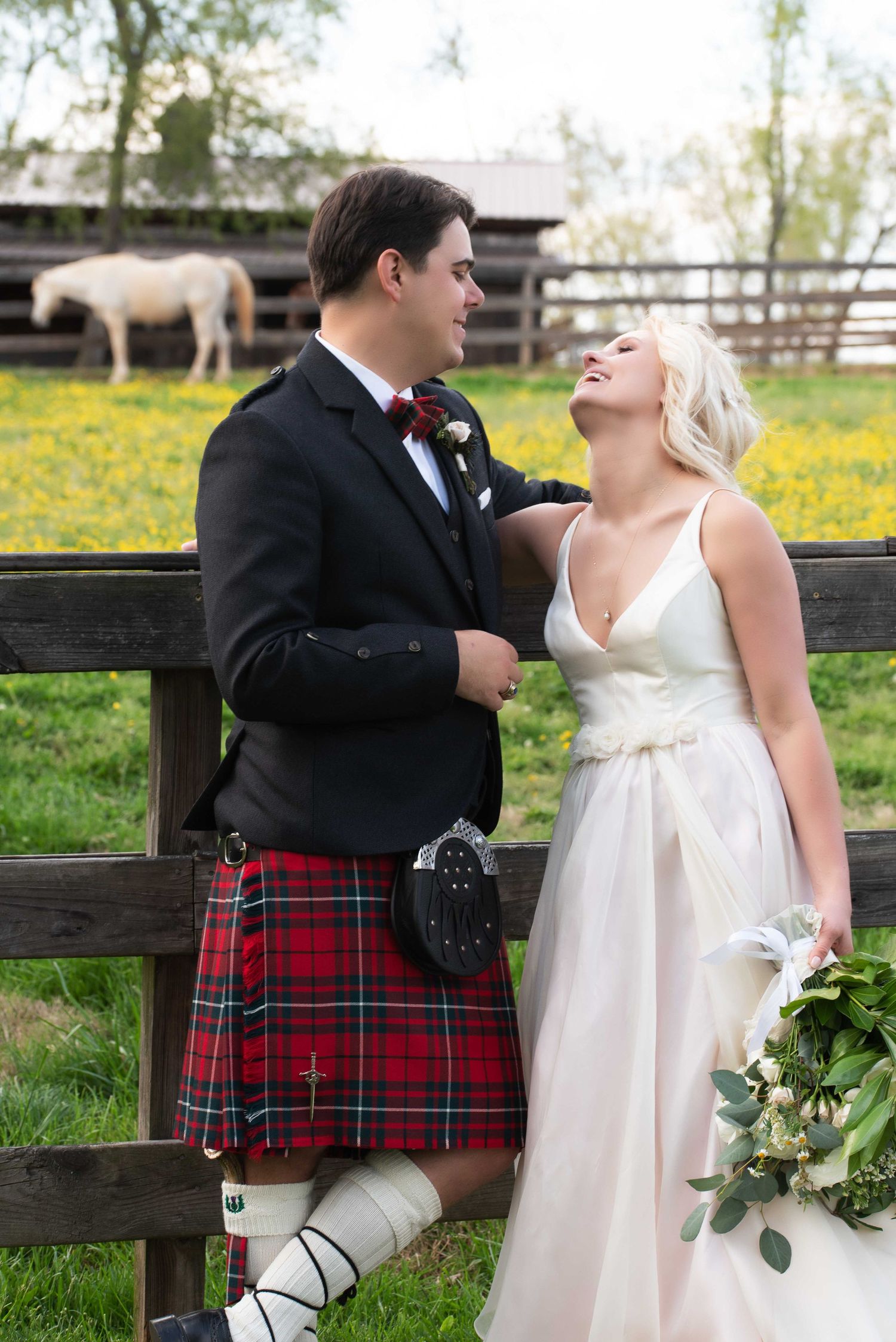Bride in white gown and groom in Scottish kilt embrace by a wooden fence with a white horse grazing in background.