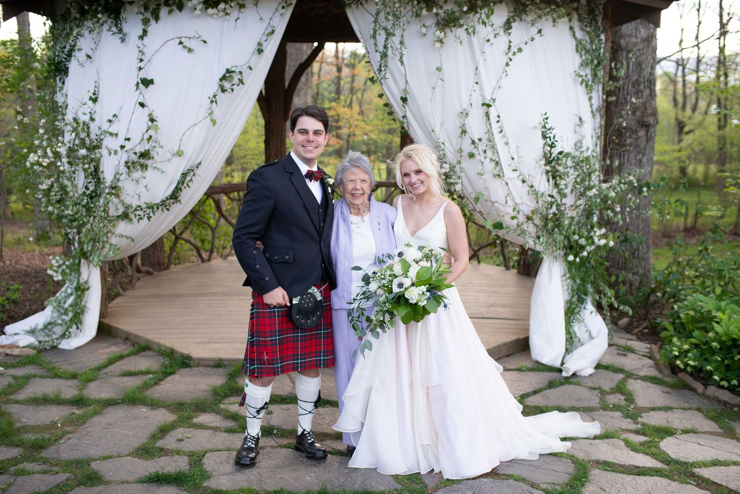 Bride in white gown and groom in Scottish tartan kilt pose with officiant under floral wedding arch outdoors.
