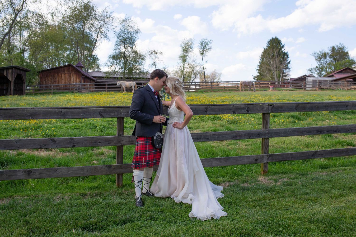Bride and groom kissing by a wooden fence at a rustic farm wedding, groom wearing a red tartan kilt.
