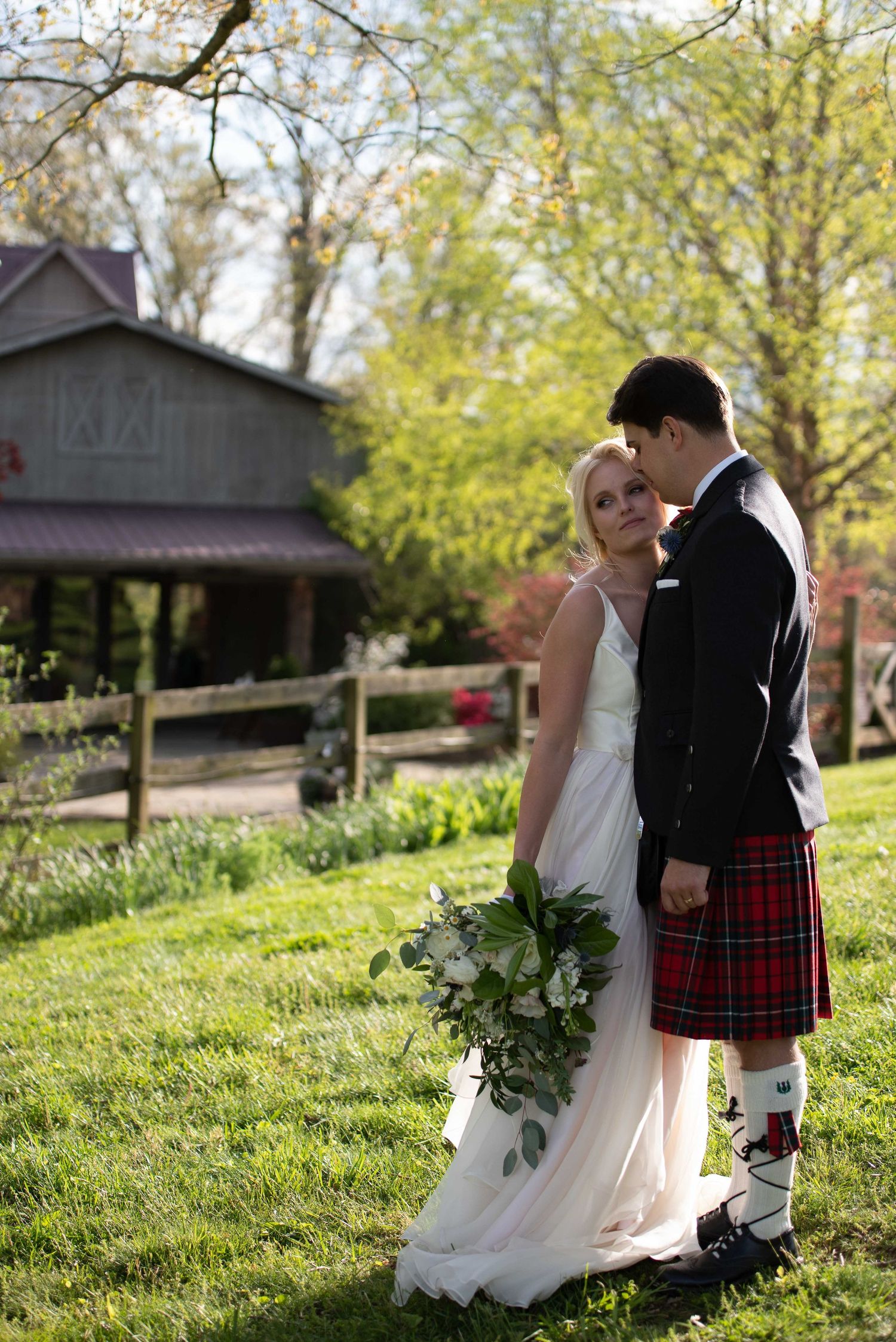 Bride in white gown and groom in red tartan kilt embrace outdoors near rustic barn on lush green wedding grounds.