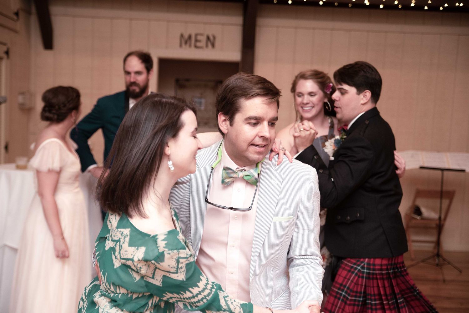 Couple dancing at wedding reception, man in bow tie and woman in green dress, guests in background near men's restroom.