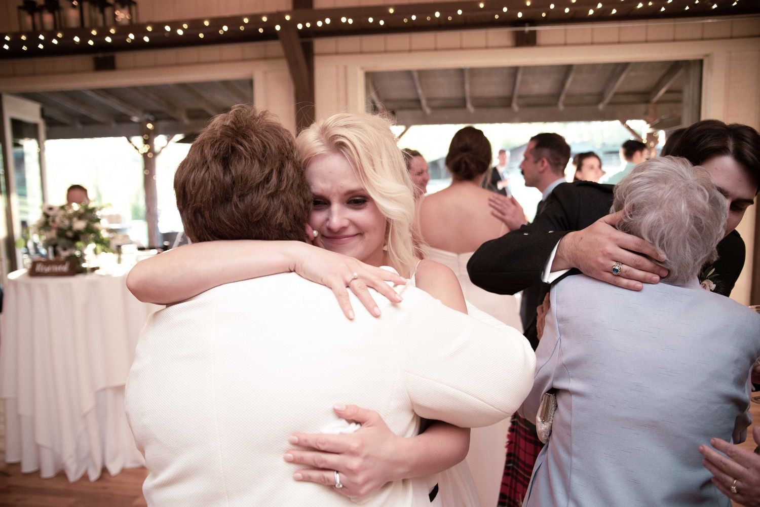 Bride in white dress sharing a warm hug with guests at an indoor wedding reception venue.