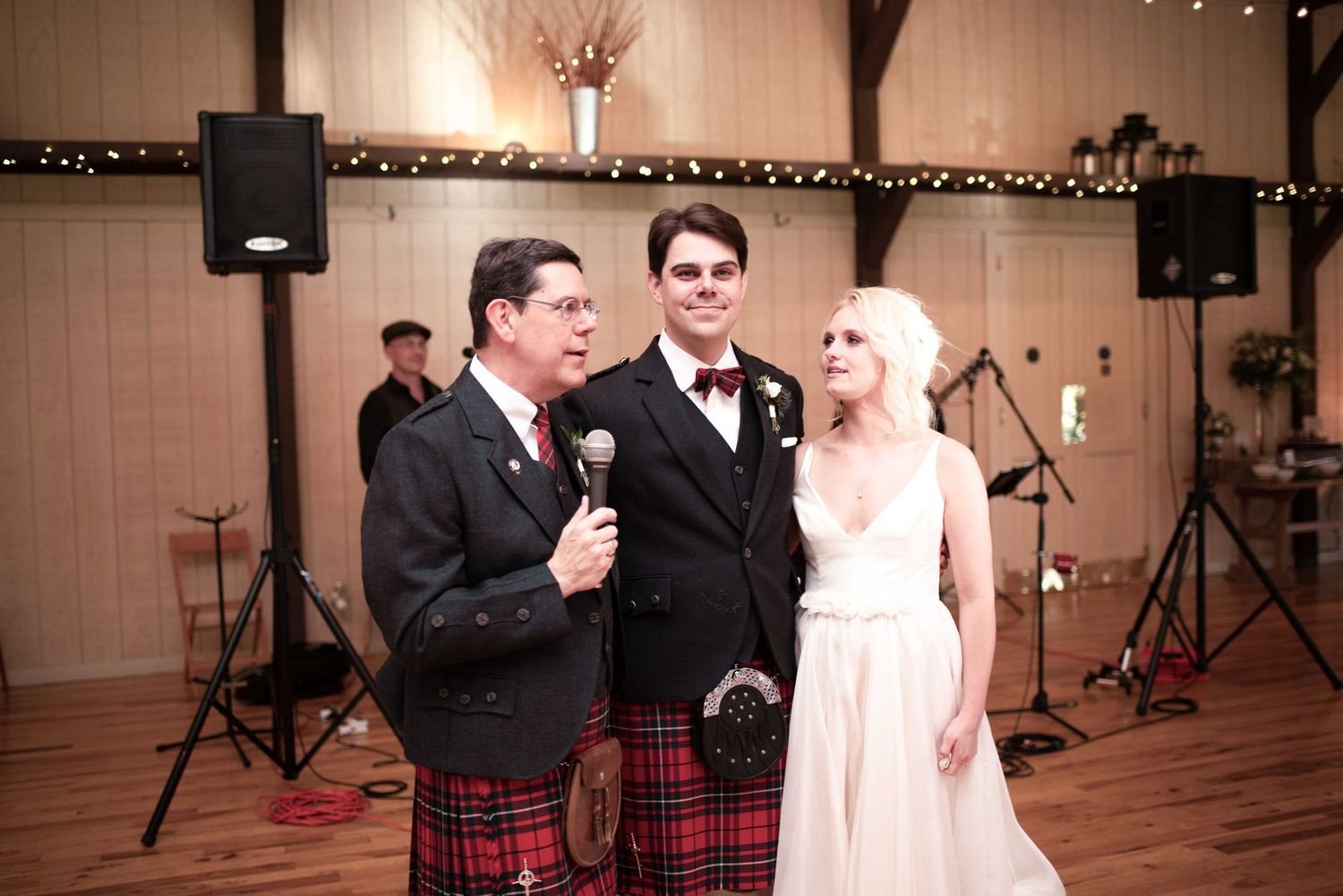 Man in Scottish kilt speaks at wedding reception with groom in bow tie and bride in white gown at rustic venue.