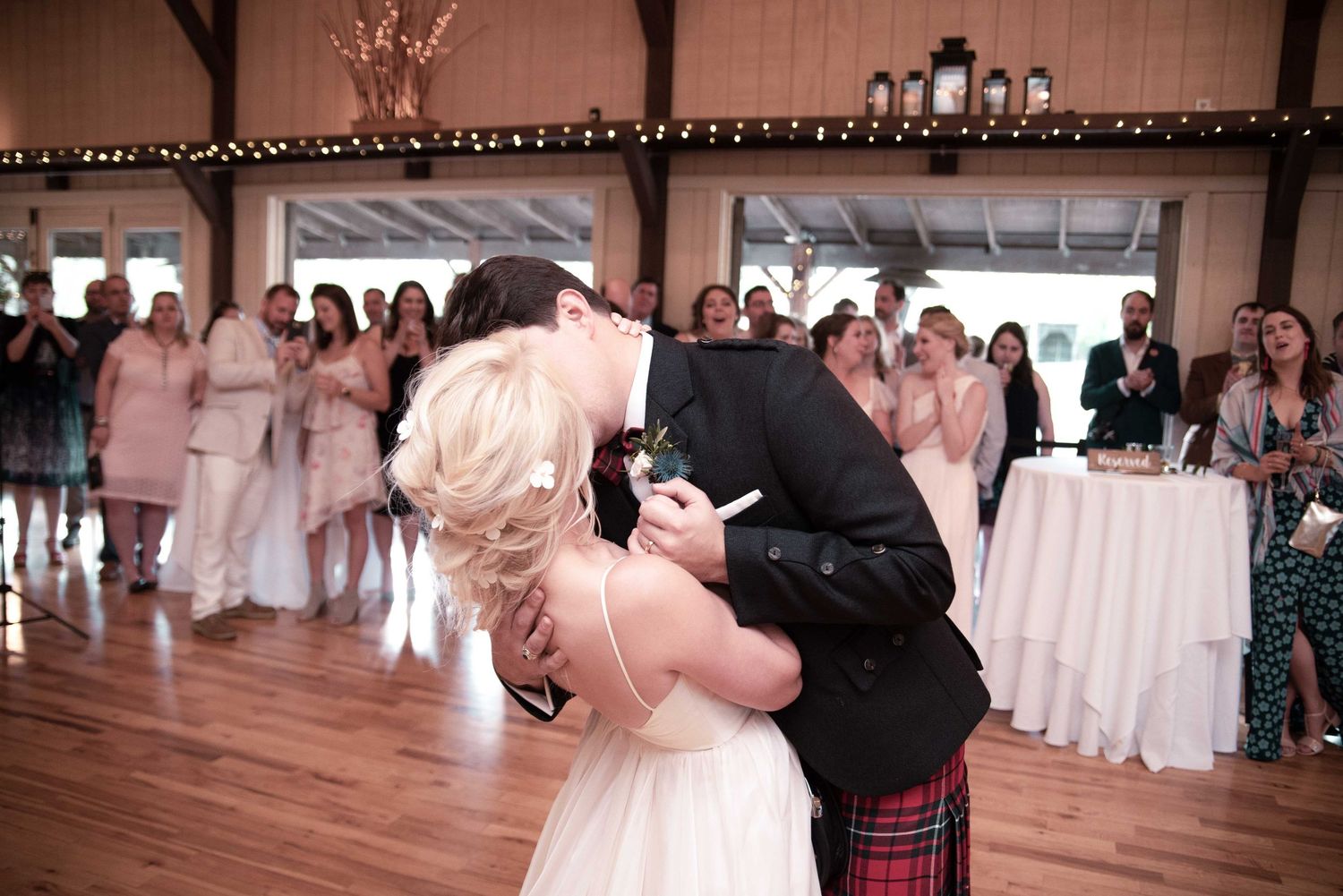 Bride and groom share first dance kiss at rustic wedding reception, groom wearing traditional Scottish kilt.
