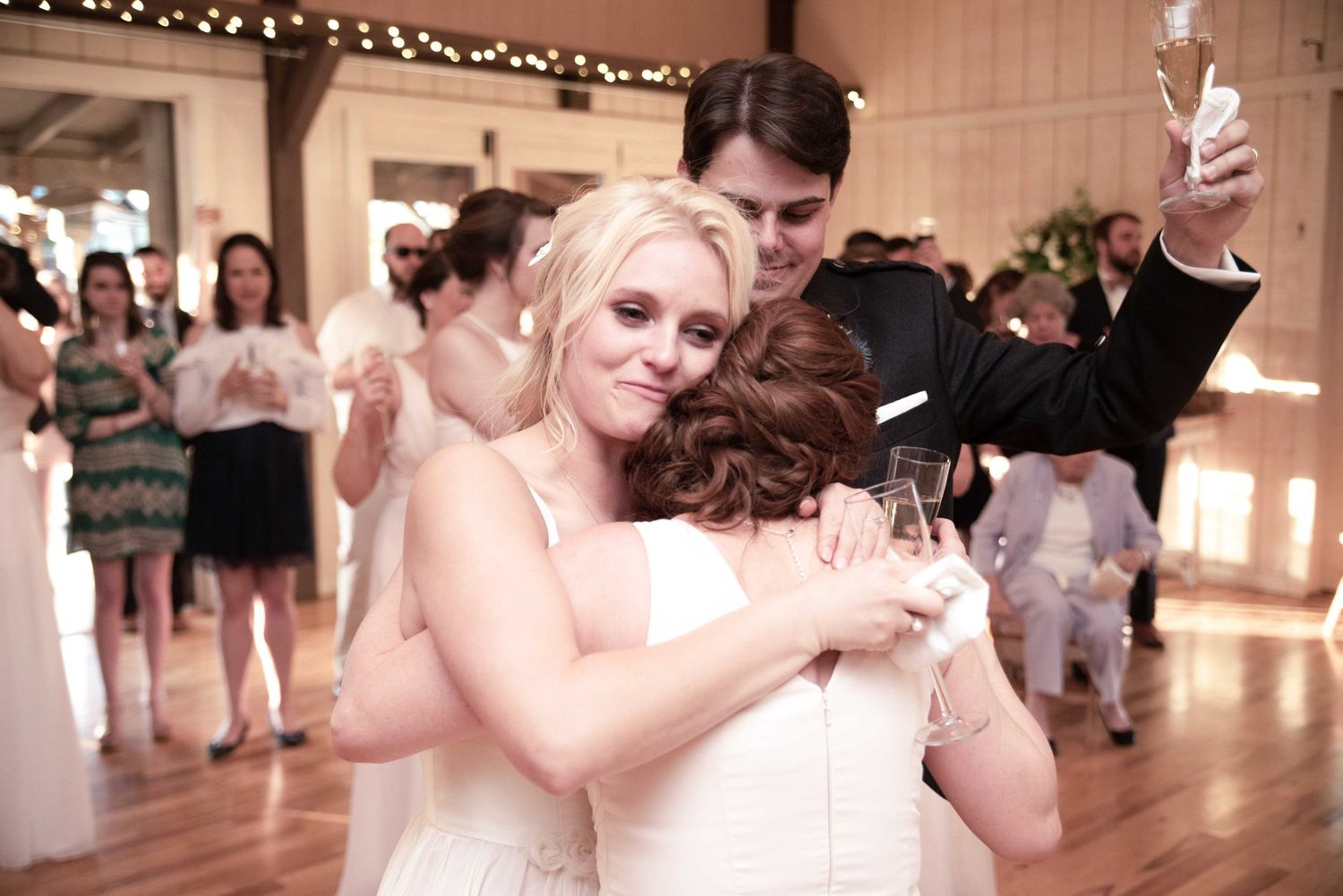 Bride hugging guest at wedding reception while groom raises champagne glass behind her.