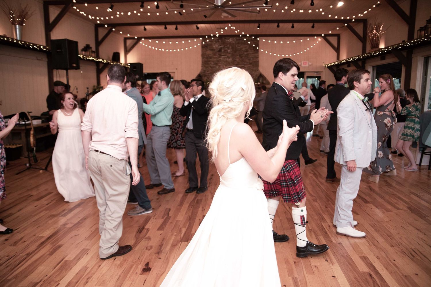 Wedding reception guests dancing in a rustic barn venue with string lights, bride in white gown and groom in red tartan kilt.