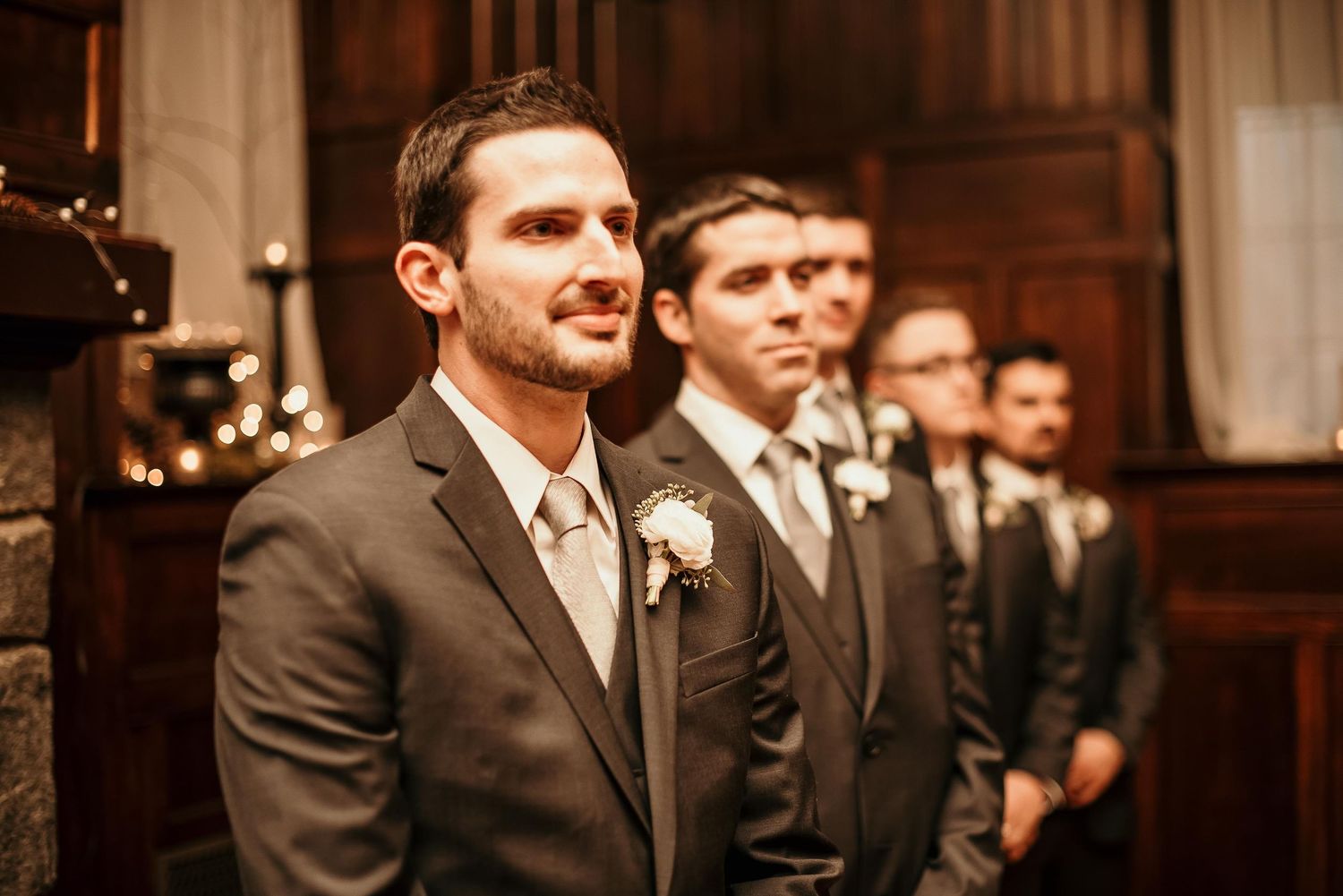 Groomsmen in gray suits with boutonnieres stand at wedding altar, groom in foreground looking emotional.